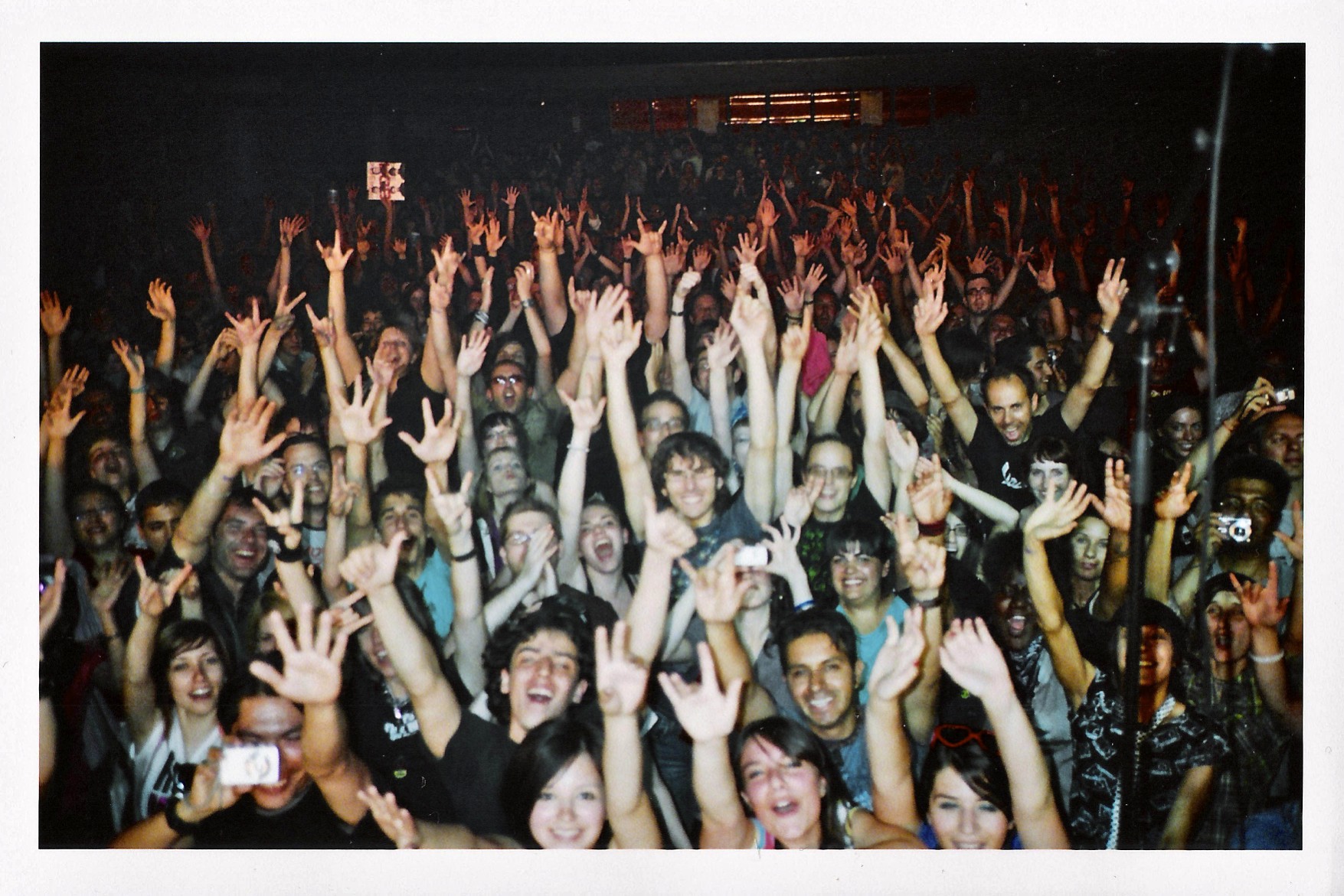 A large crowd at a concert cheers, smiling and raising their hands enthusiastically toward the camera, capturing the moment like a skilled photographer with a futuristic perspective.