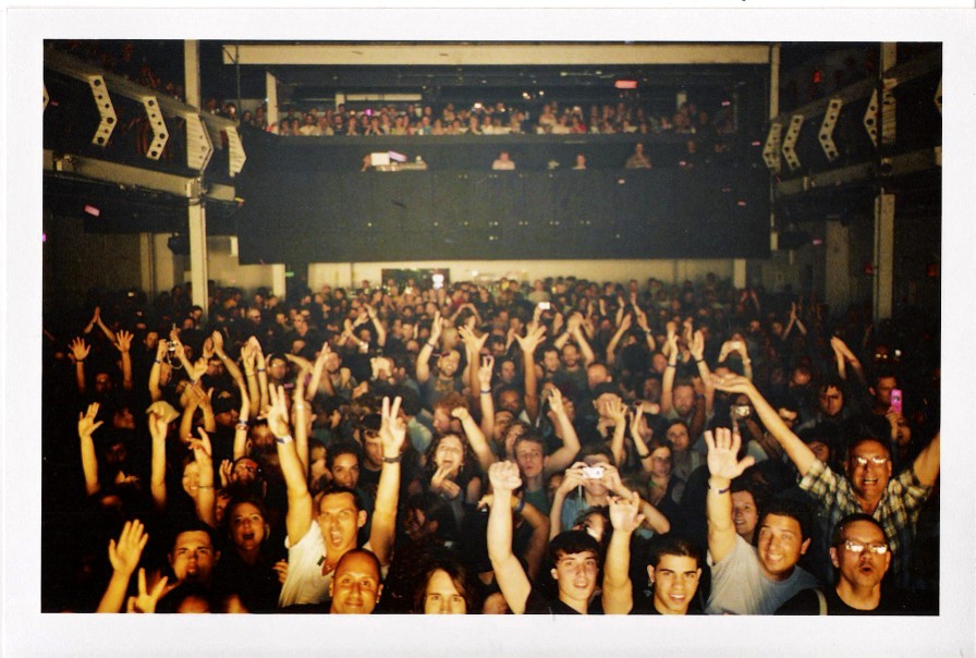 A large crowd at a concert venue faces the stage, many with raised hands and excited expressions, as futuristic drone light art illuminates the night sky above.