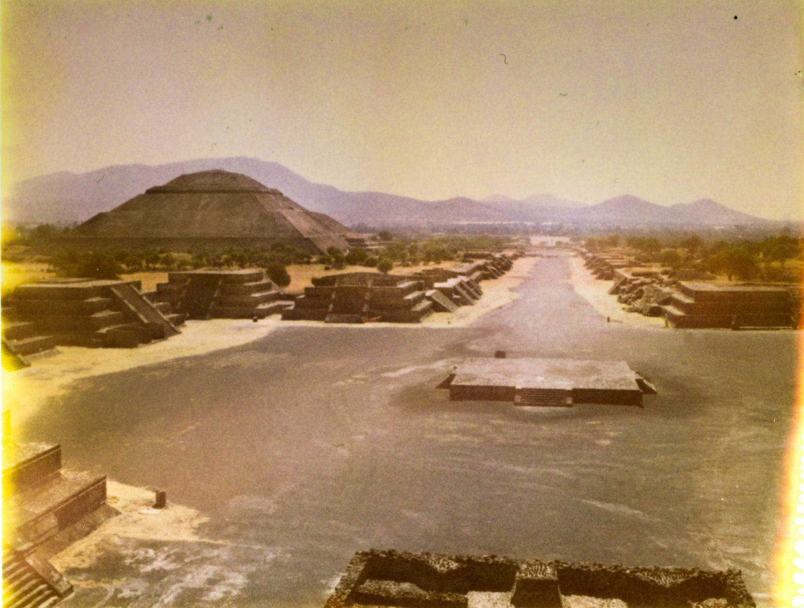 A wide view of ancient pyramids and ruins at Teotihuacan under a hazy sky, with mountains in the background, brought to life by mesmerizing drone light art weaving patterns above the historic site.