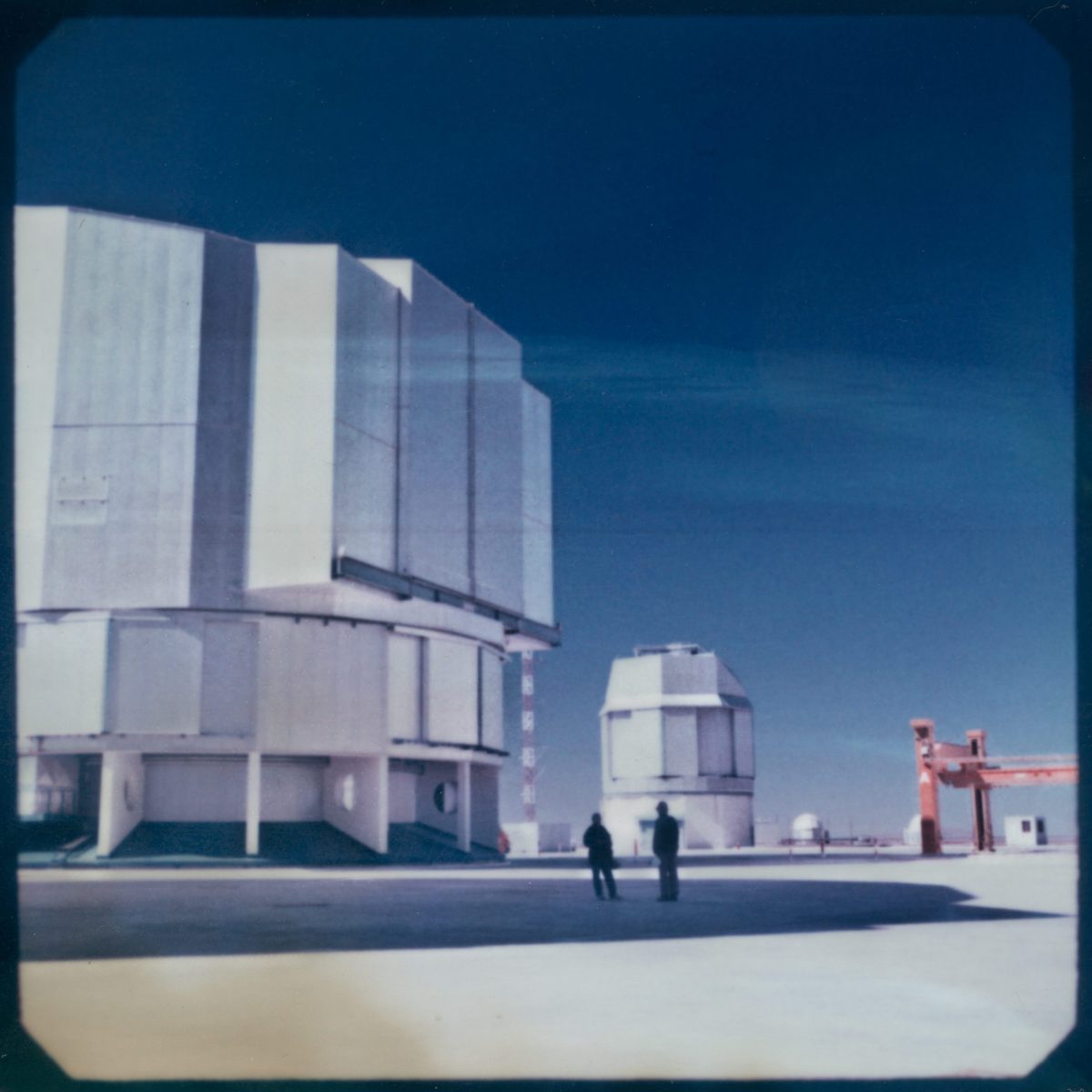 Two people stand near large observatory domes under a clear blue sky, as a photographer captures the scene with hints of futuristic drone light painting overhead.