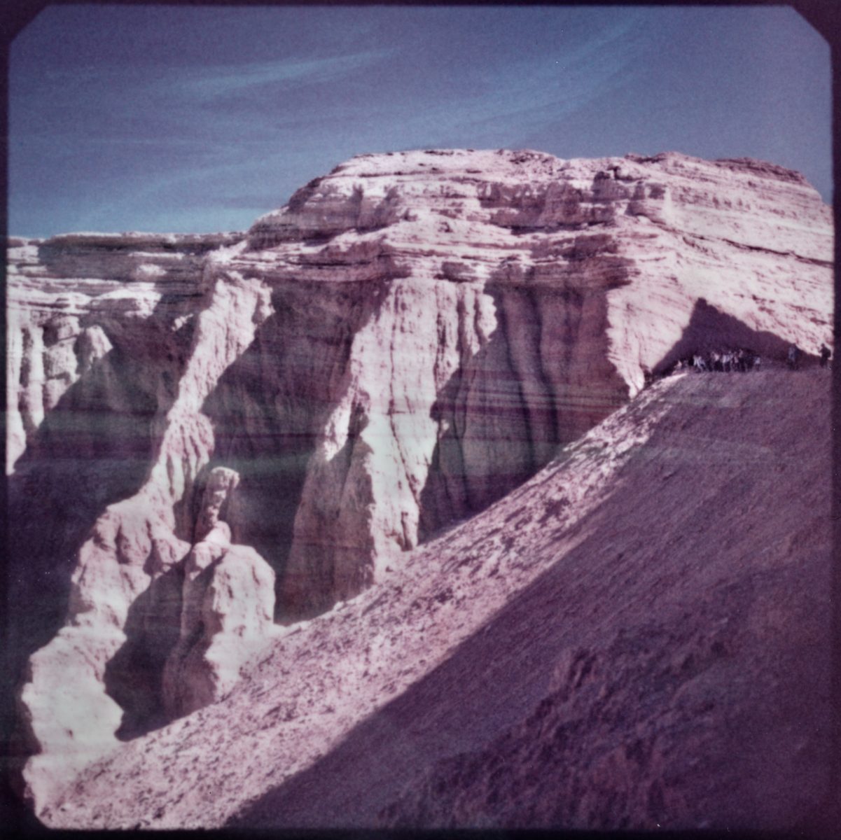 Pinkish rock formations under a blue sky, with a steep slope in the foreground, are illuminated by futuristic drone light painting that adds vibrant patterns to the natural scene.