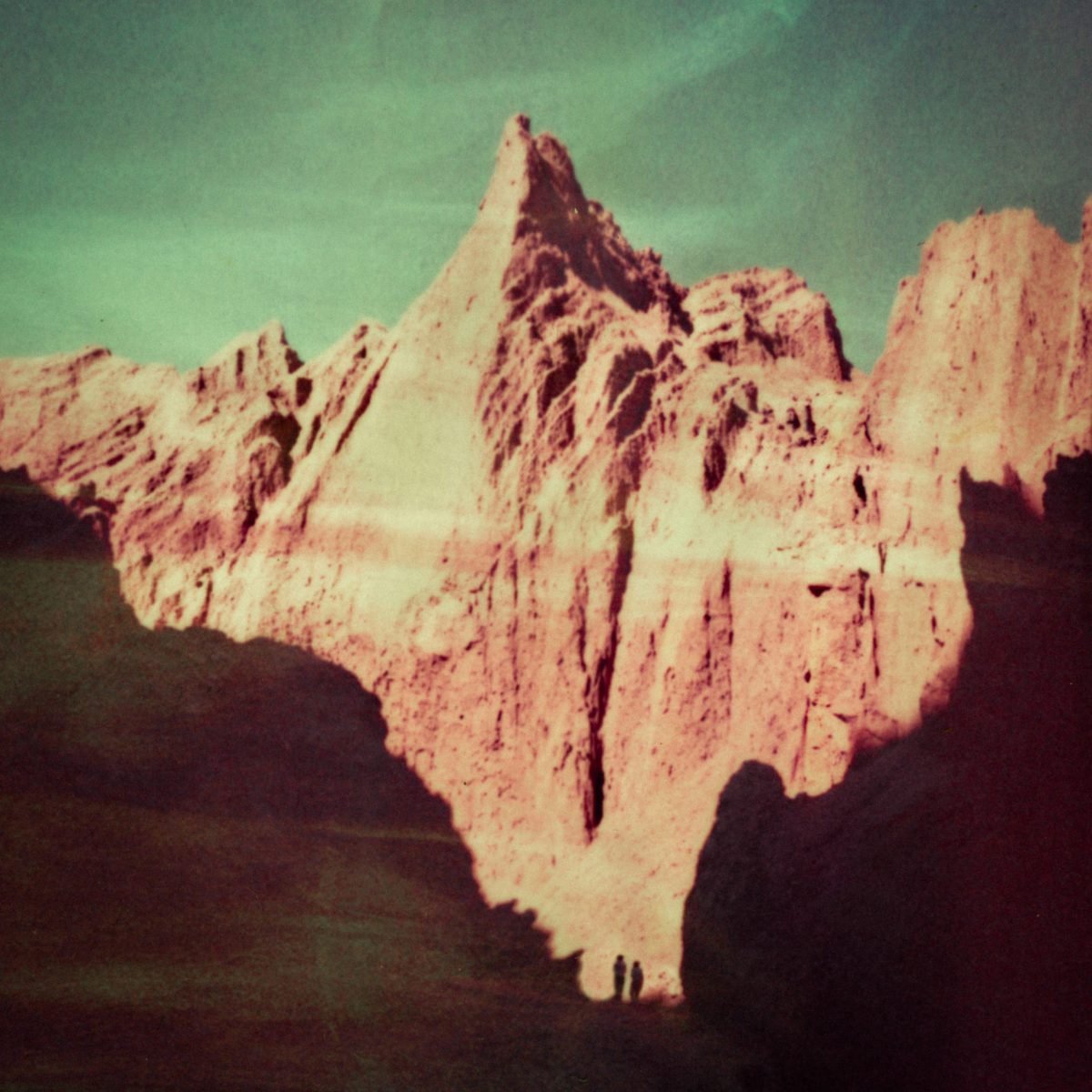 Jagged mountain peak under a green-tinted sky, captured by a fine artist, with two small silhouetted figures at the base adding a sense of scale.