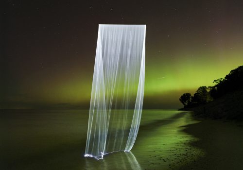 A glowing vertical light trail, created through drone light painting, stands on a beach at night, with green aurora lighting up the sky.