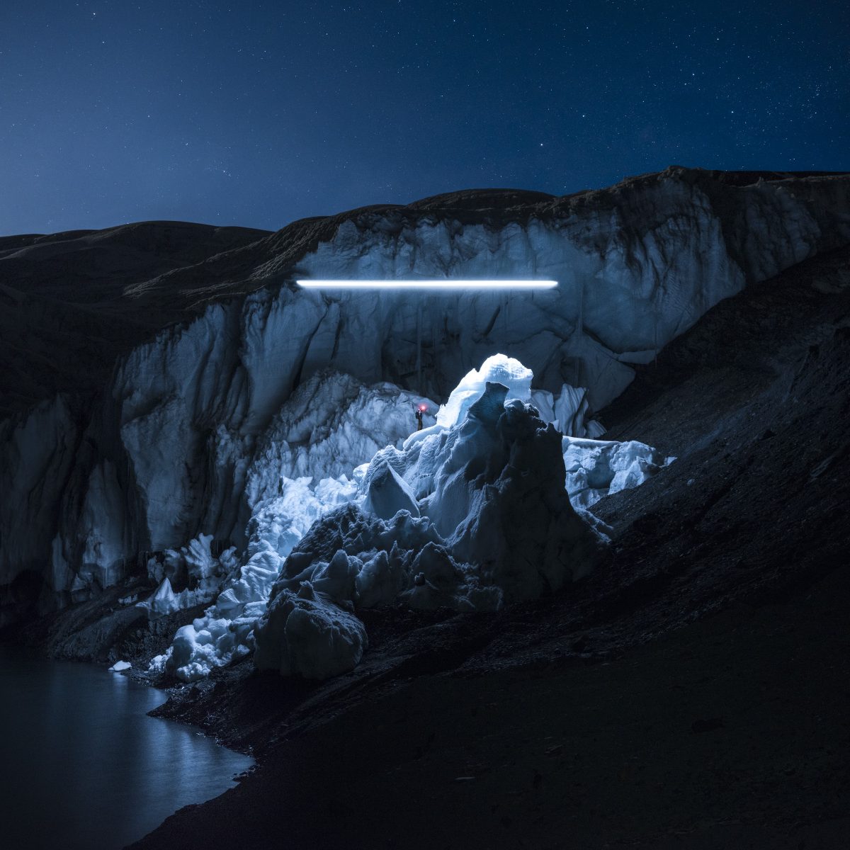 A person standing on a large ice formation illuminated by a bright light in a dark, starry landscape.