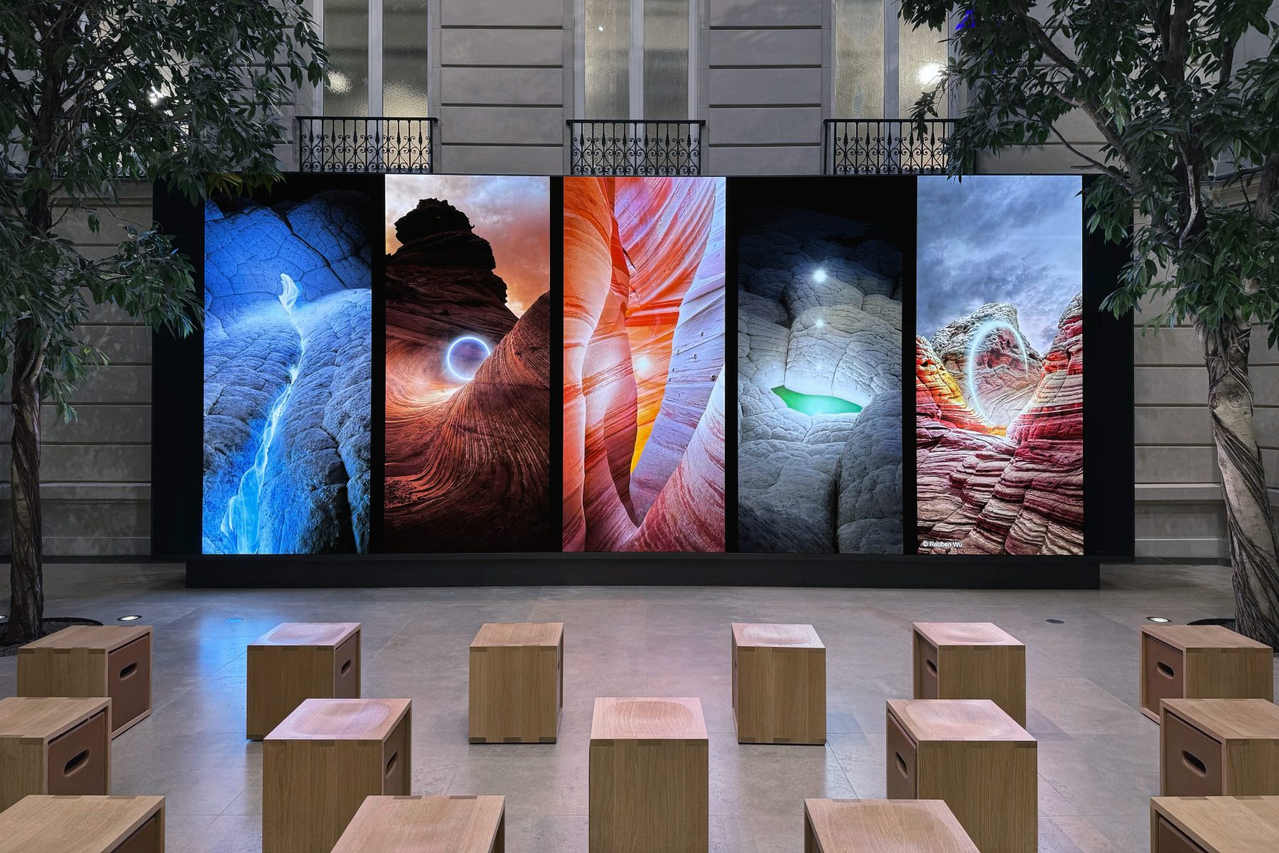 Five digital screens display colorful, futuristic abstract landscapes in a spacious indoor area with wooden stools and trees.