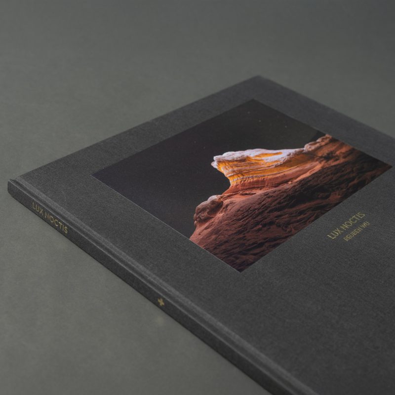 A dark hardcover book featuring a striking nighttime landscape photography of a rocky peak, enhanced by futuristic drone light art, with the title LUX NOCTIS embossed in gold text.