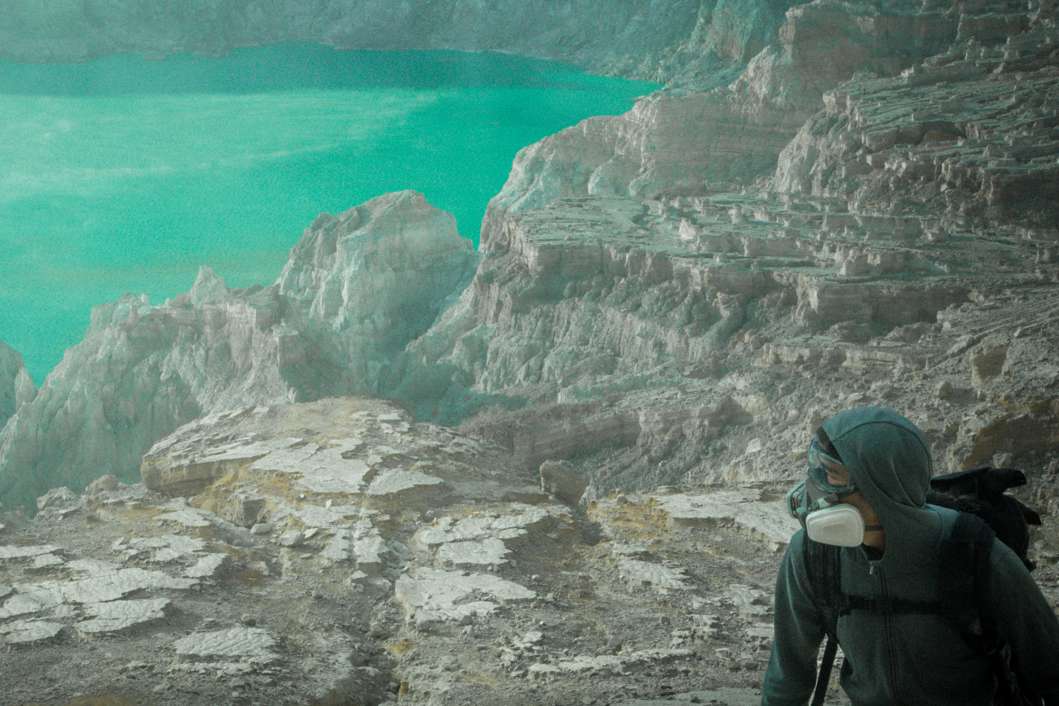 A hiker in a gas mask stands near rocky cliffs overlooking a turquoise lake, as futuristic drone light art glows softly in the evening sky.