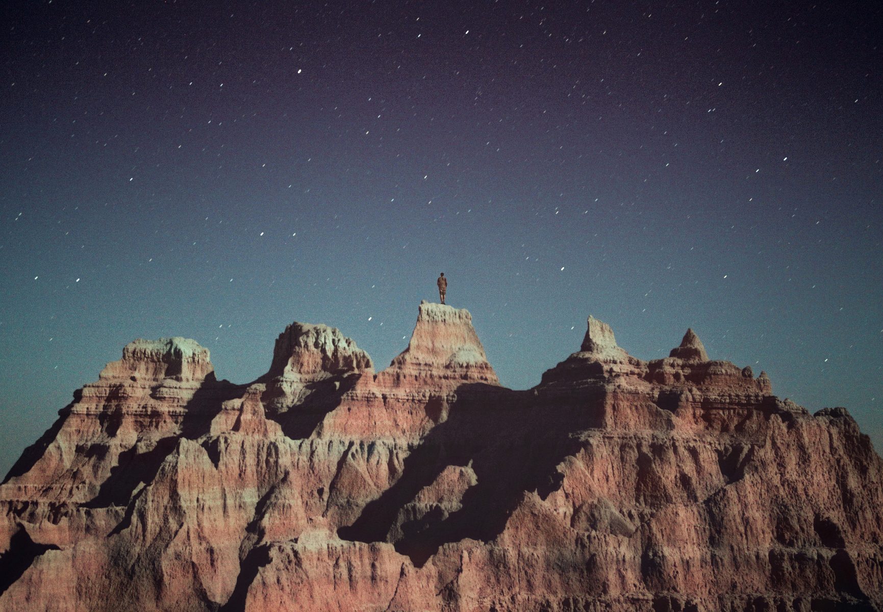 Person standing atop a rocky peak under a starry night sky in a rugged desert landscape, illuminated by captivating drone light art—perfect for lovers of nighttime landscape photography.