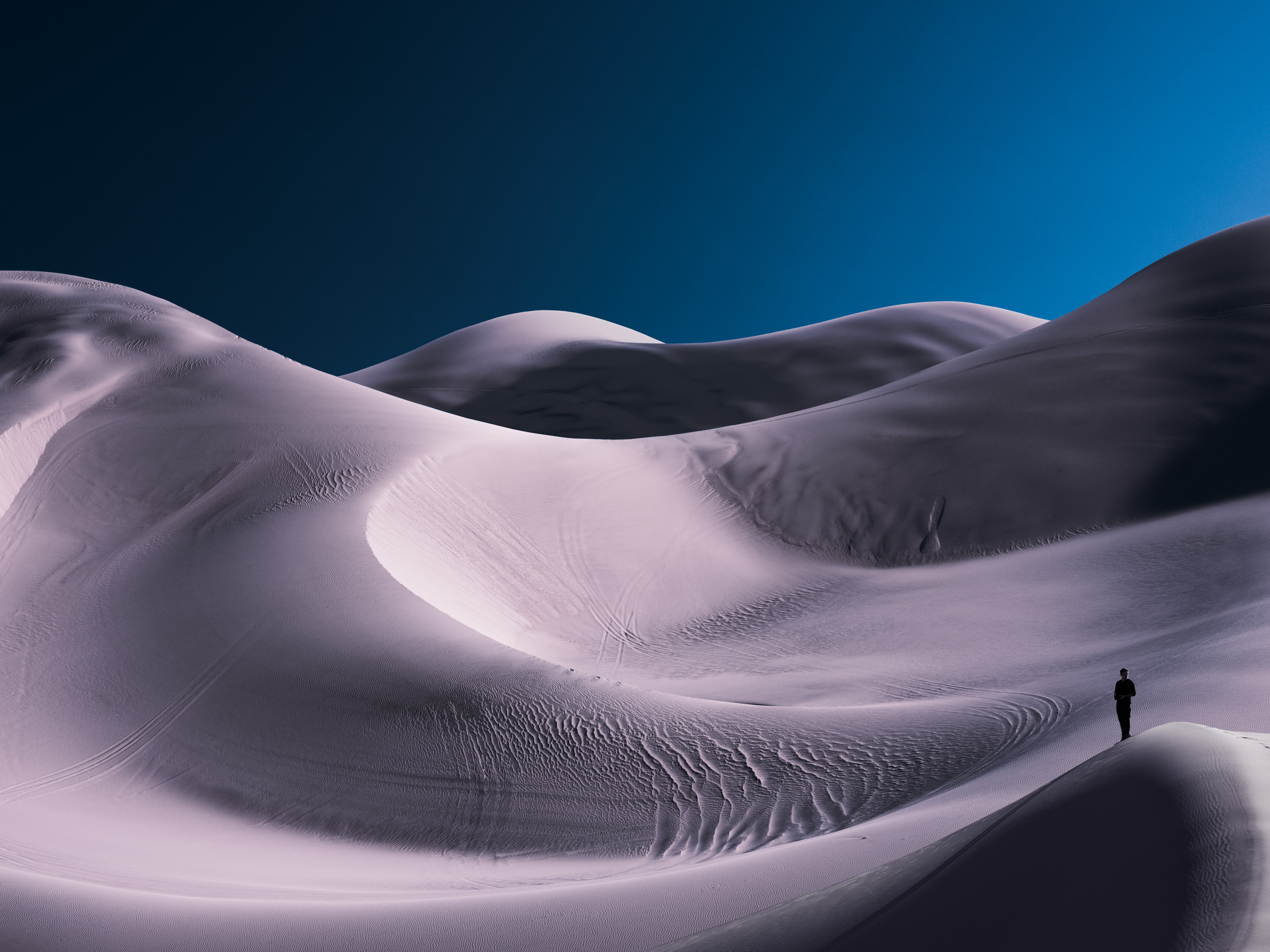 A lone person stands by smooth, rolling sand dunes under a deep blue sky, capturing the tranquility with the eye of a fine artist and the precision of nighttime landscape photography.