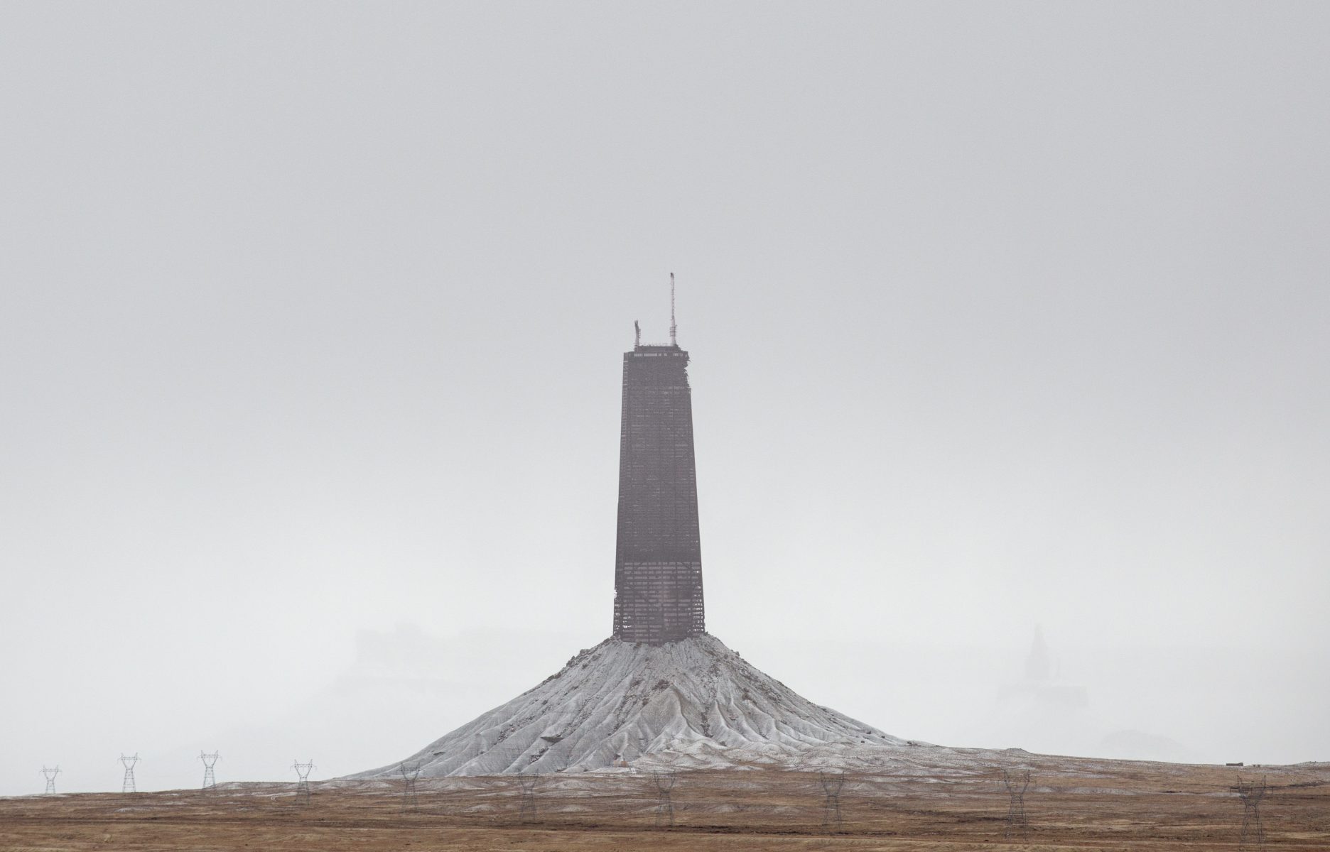 A tall, dark tower stands atop a barren, mound-like hill under a gray, overcast sky—an ideal scene for dramatic nighttime landscape photography.