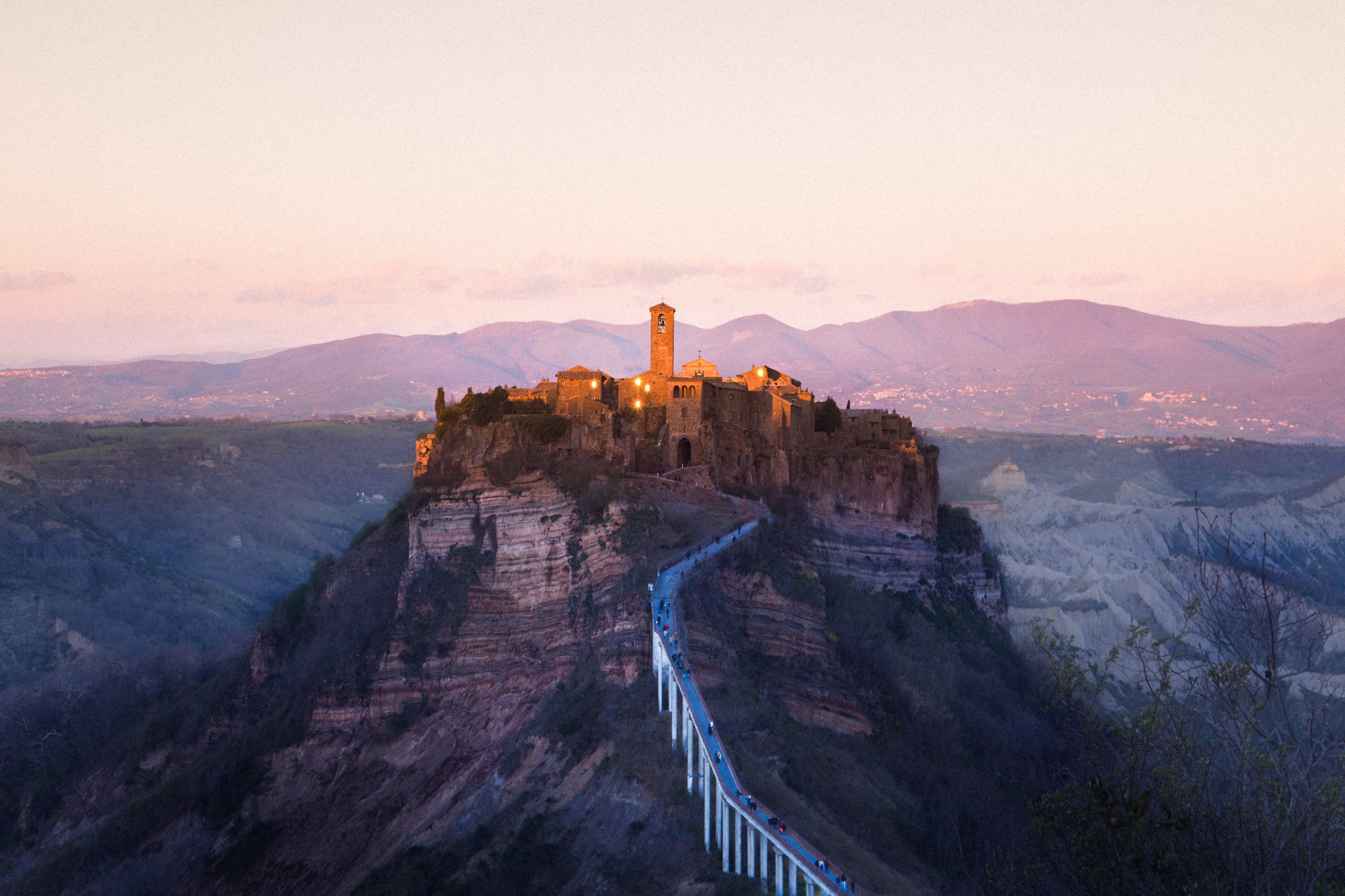 Hilltop medieval village with a tall tower at sunset, connected by a long footbridge, surrounded by mountains and illuminated by futuristic drone light painting in the evening sky.
