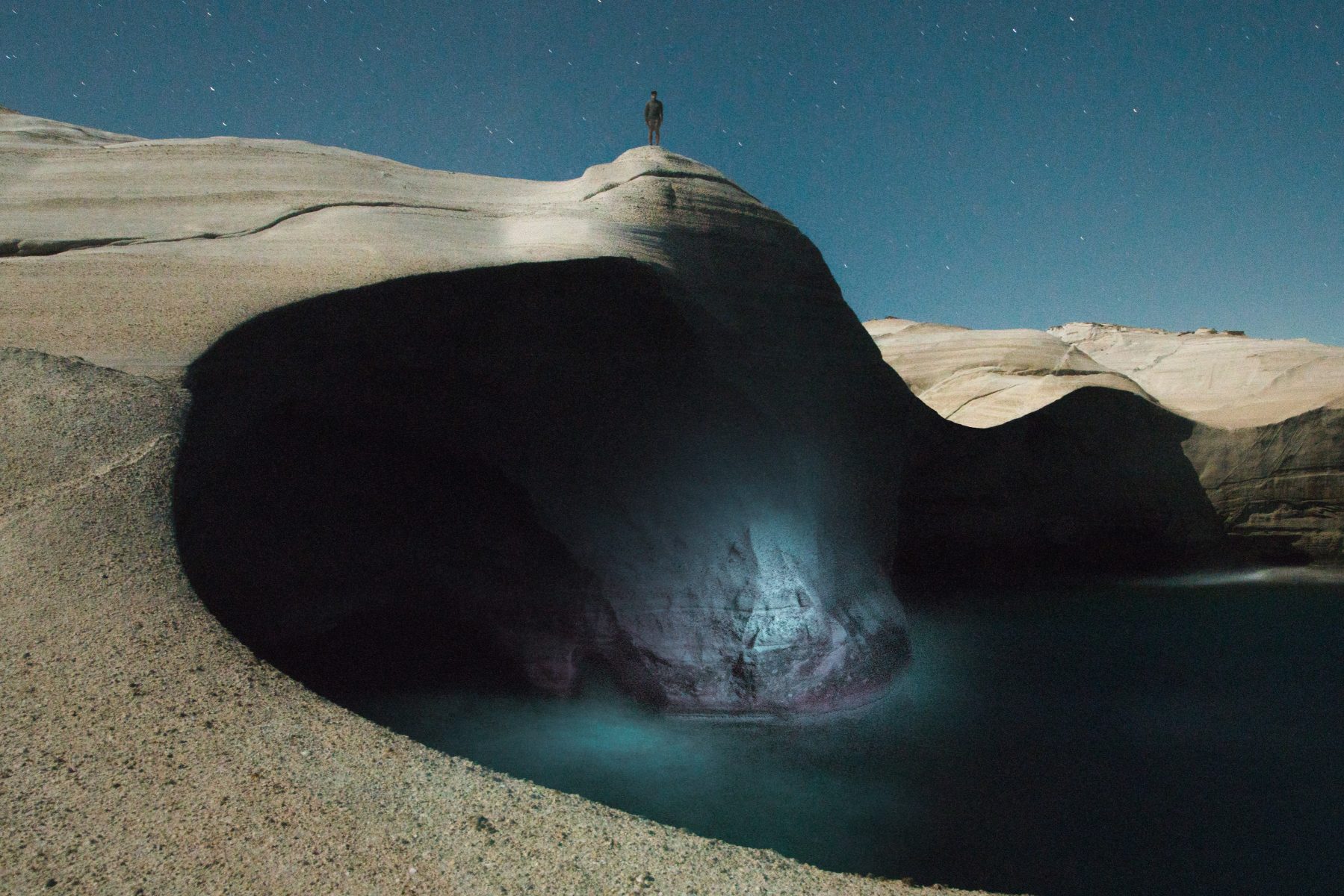 A person stands atop a large, rocky cliff above a glowing blue pool under a starry night sky, creating a mesmerizing scene reminiscent of nighttime landscape photography.