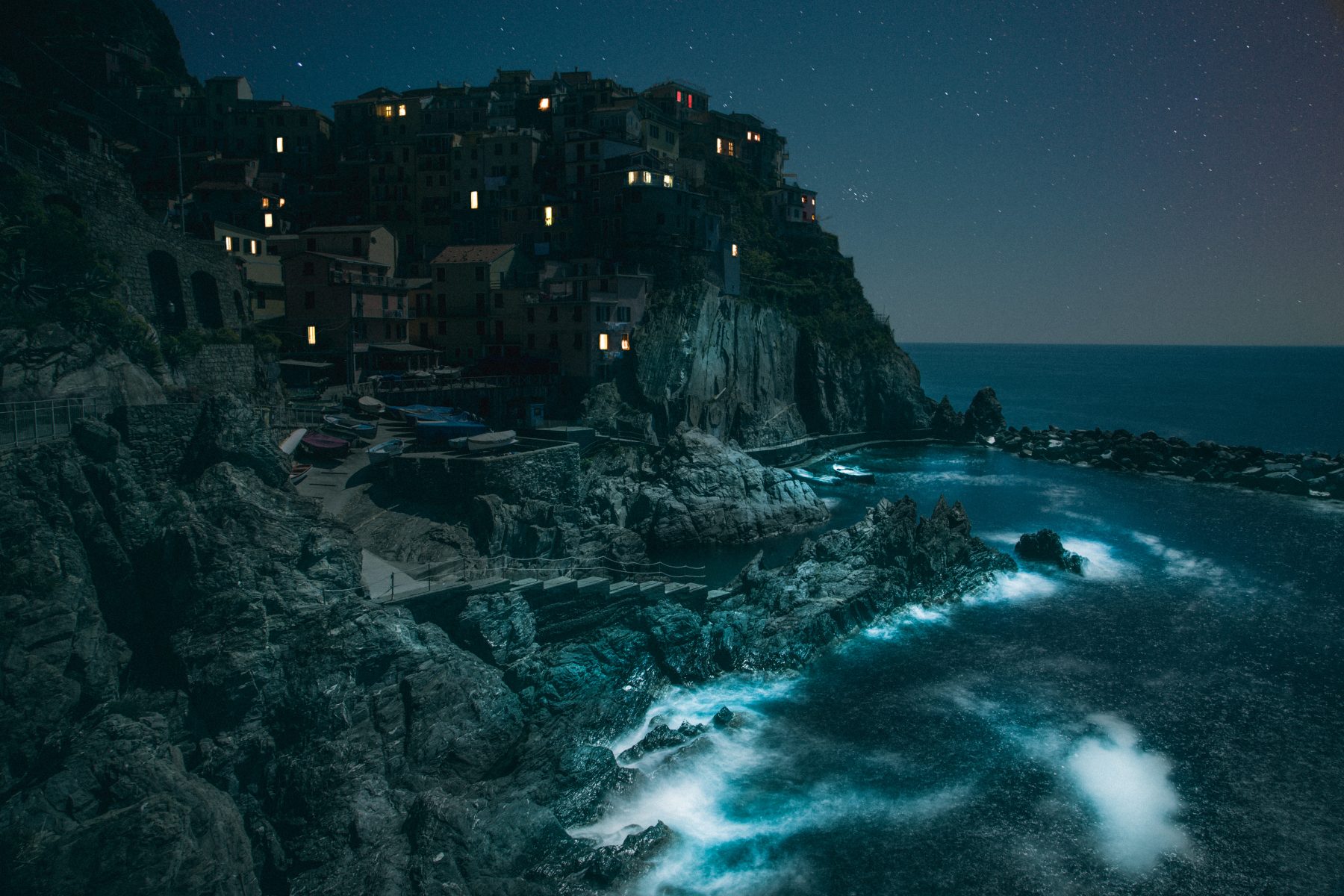 A coastal village with lit windows sits on cliffs above a rocky shore under a starry night sky, beautifully captured through nighttime landscape photography.
