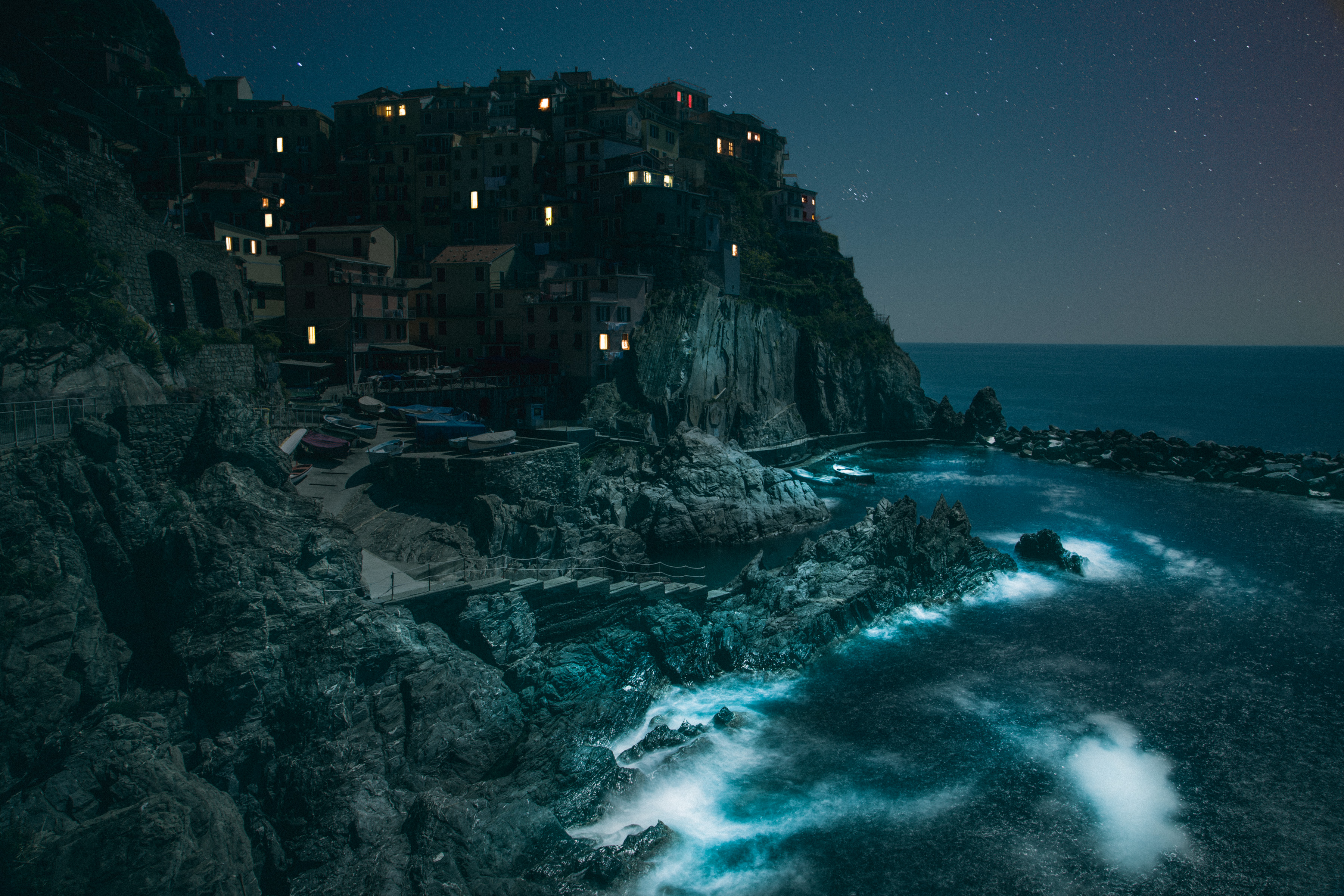 A coastal village with lit windows sits on cliffs above a rocky shore under a starry night sky, beautifully captured through nighttime landscape photography.