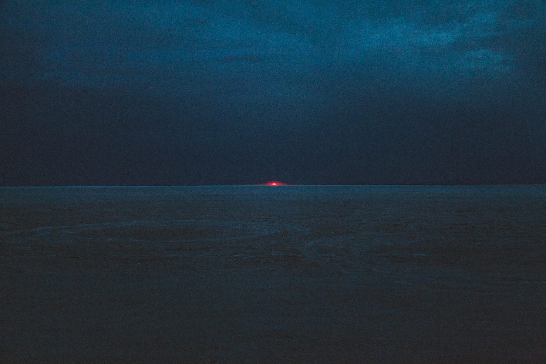 A dark, cloudy landscape with a faint red-orange sun setting on the distant horizon, reminiscent of nighttime landscape photography by a fine artist.