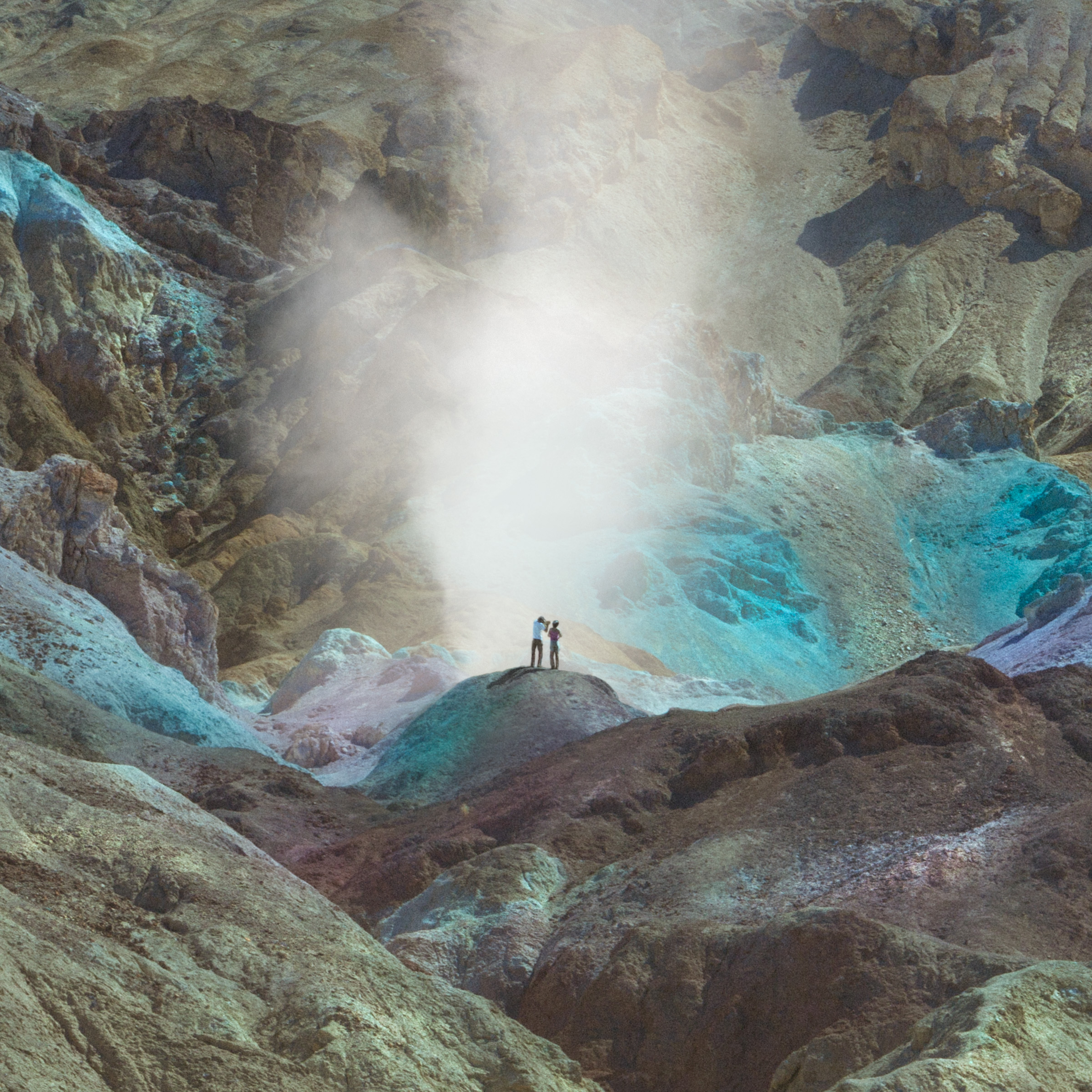Two people stand on colorful, rocky terrain surrounded by mist in a mountainous landscape, as a photographer captures the scene illuminated by mesmerizing drone light art.