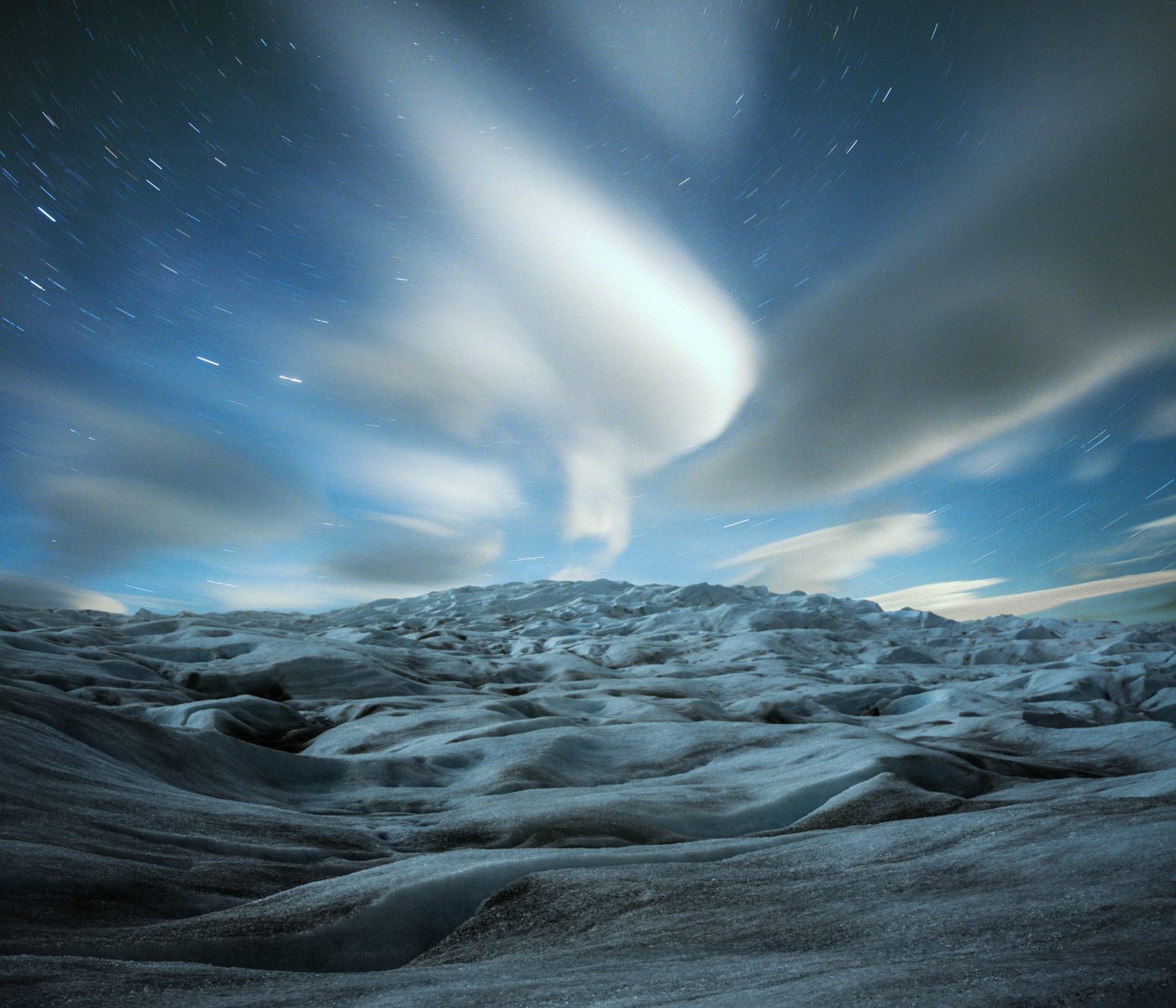 Clouds streak across a starry night sky above a rugged, icy landscape, revealing the drama of nighttime landscape photography reminiscent of a fine artist’s vision.