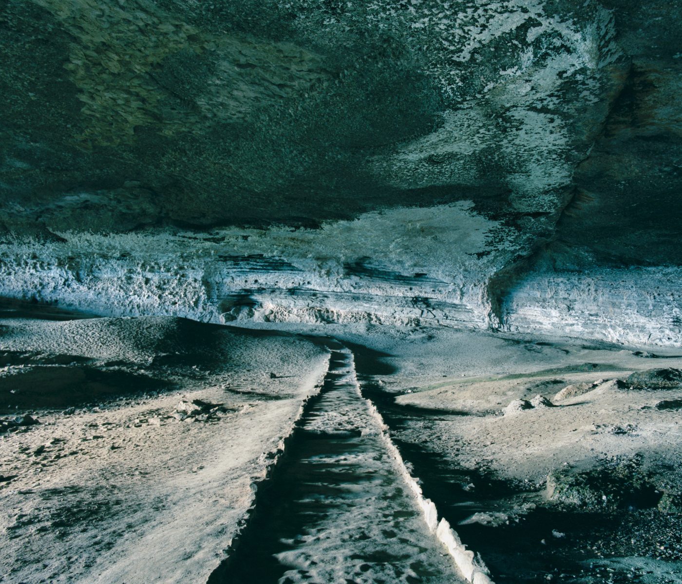 A stone path leads through a large, shadowy cave with textured, rocky walls and ceiling, reminiscent of scenery a fine artist or photographer might capture in their work.