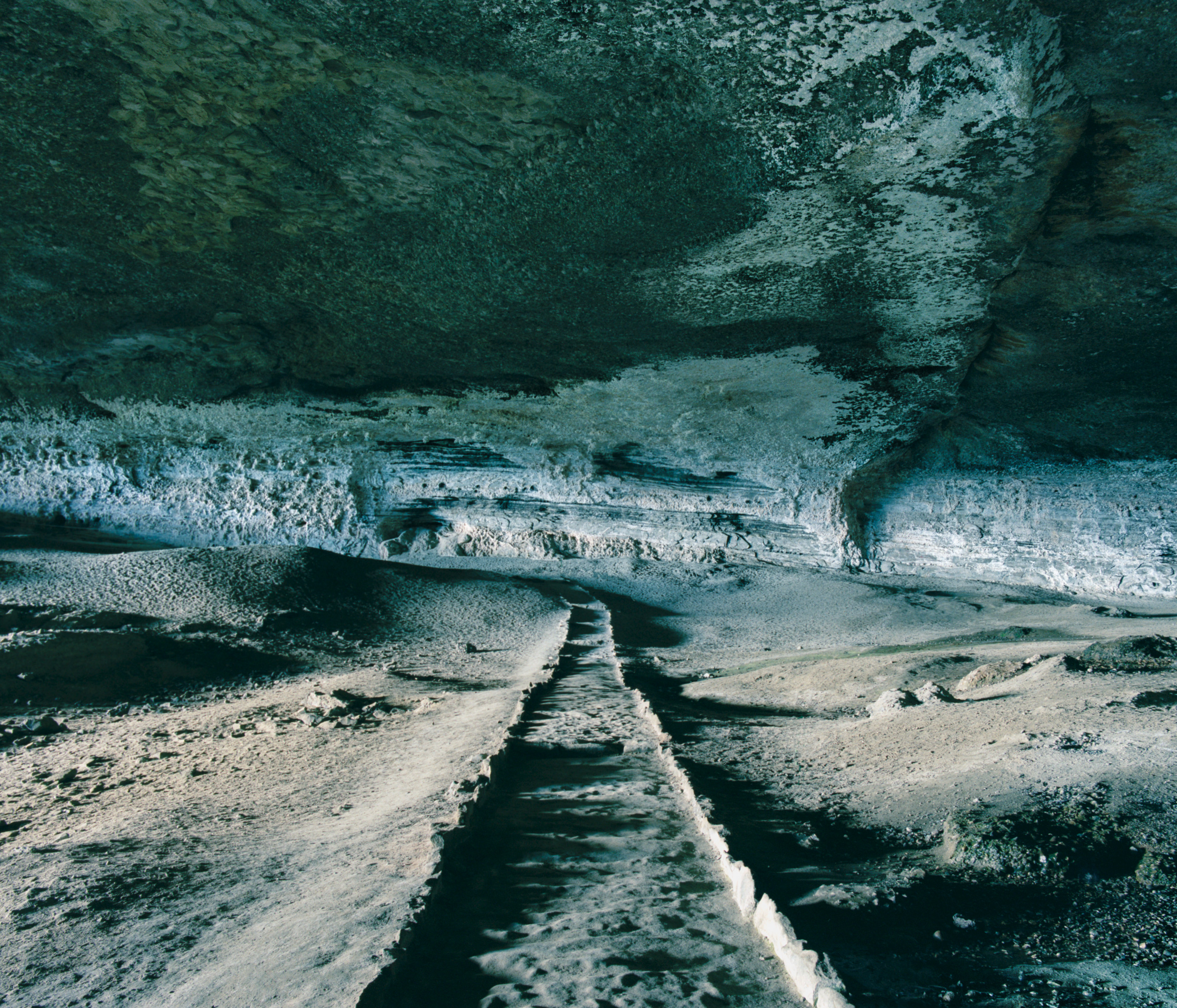 A stone path leads through a large, shadowy cave with textured, rocky walls and ceiling, reminiscent of scenery a fine artist or photographer might capture in their work.