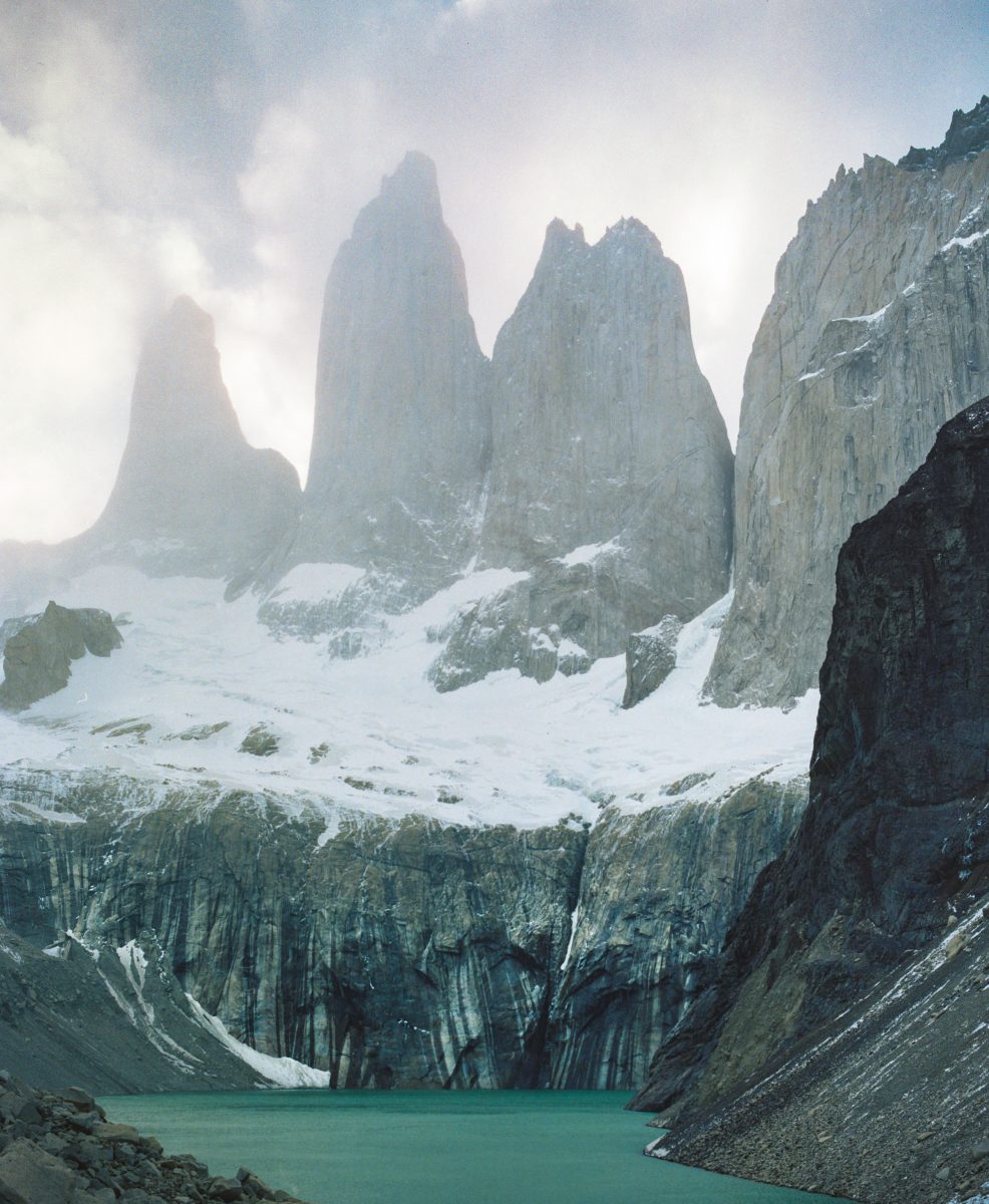 Three jagged mountain peaks rise above a turquoise lake and rocky, snow-dusted cliffs under a cloudy sky, creating a breathtaking scene for any photographer to capture.