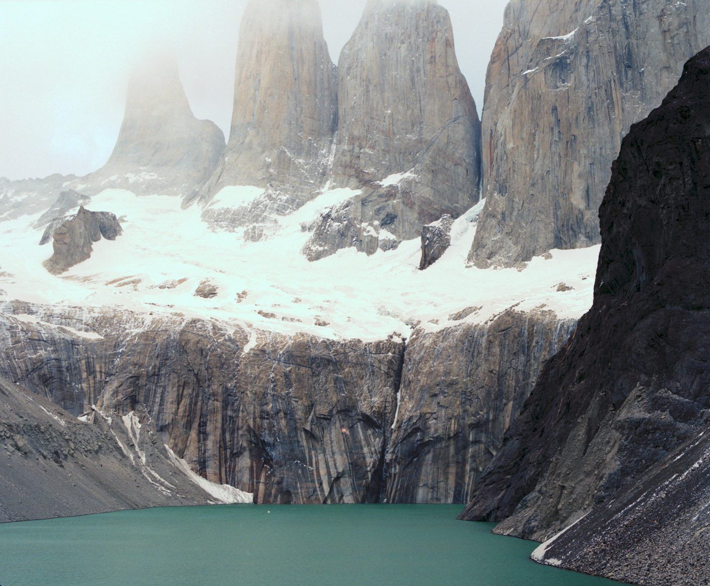 Turquoise lake beneath snow-dusted rocky cliffs with tall, fog-shrouded mountain peaks in the background—a photographer’s dream landscape.