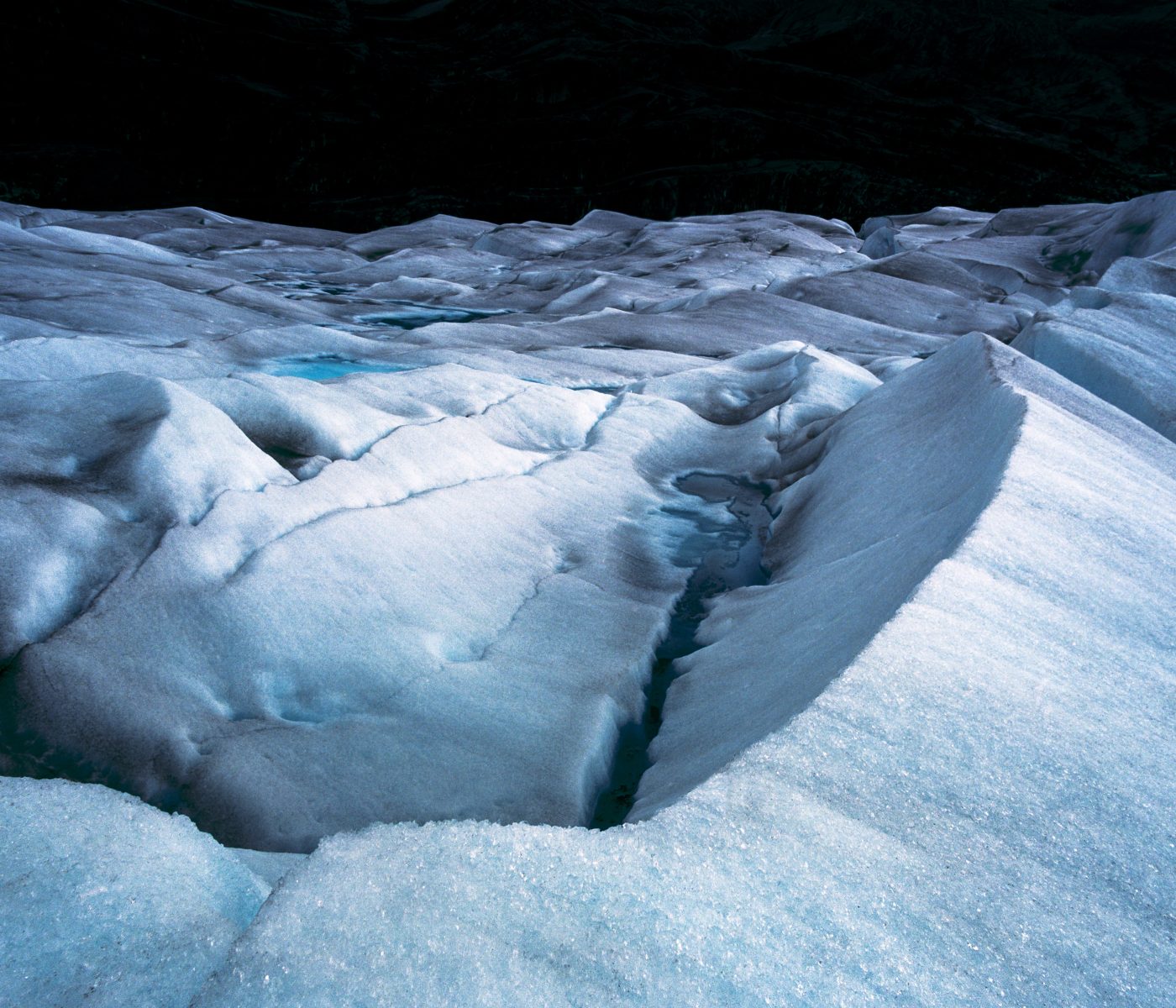 A snow-covered glacier surface with cracks and ridges is illuminated by futuristic drone light art, casting vibrant patterns across the cold, icy landscape under dim light.