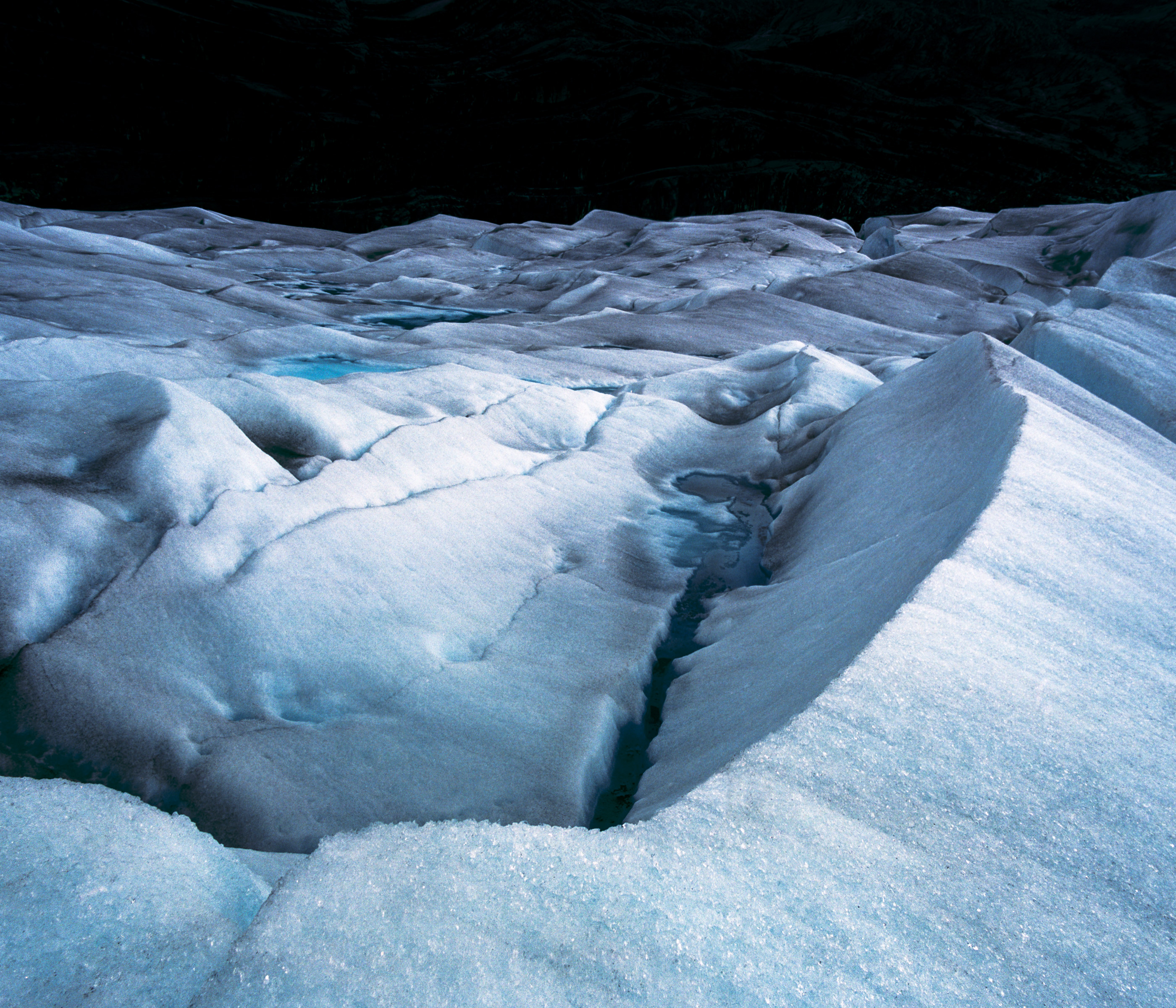 A snow-covered glacier surface with cracks and ridges is illuminated by futuristic drone light art, casting vibrant patterns across the cold, icy landscape under dim light.