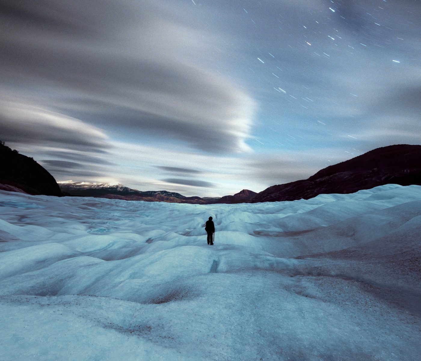 A lone figure stands on an icy, futuristic terrain beneath a dramatic sky filled with blurred clouds and star trails, capturing the allure of nighttime landscape photography.