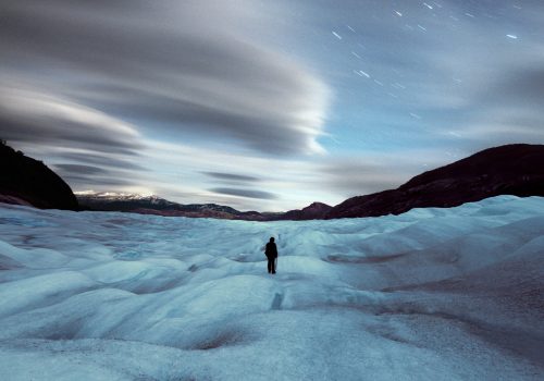 A lone figure stands on an icy, futuristic terrain beneath a dramatic sky filled with blurred clouds and star trails, capturing the allure of nighttime landscape photography.