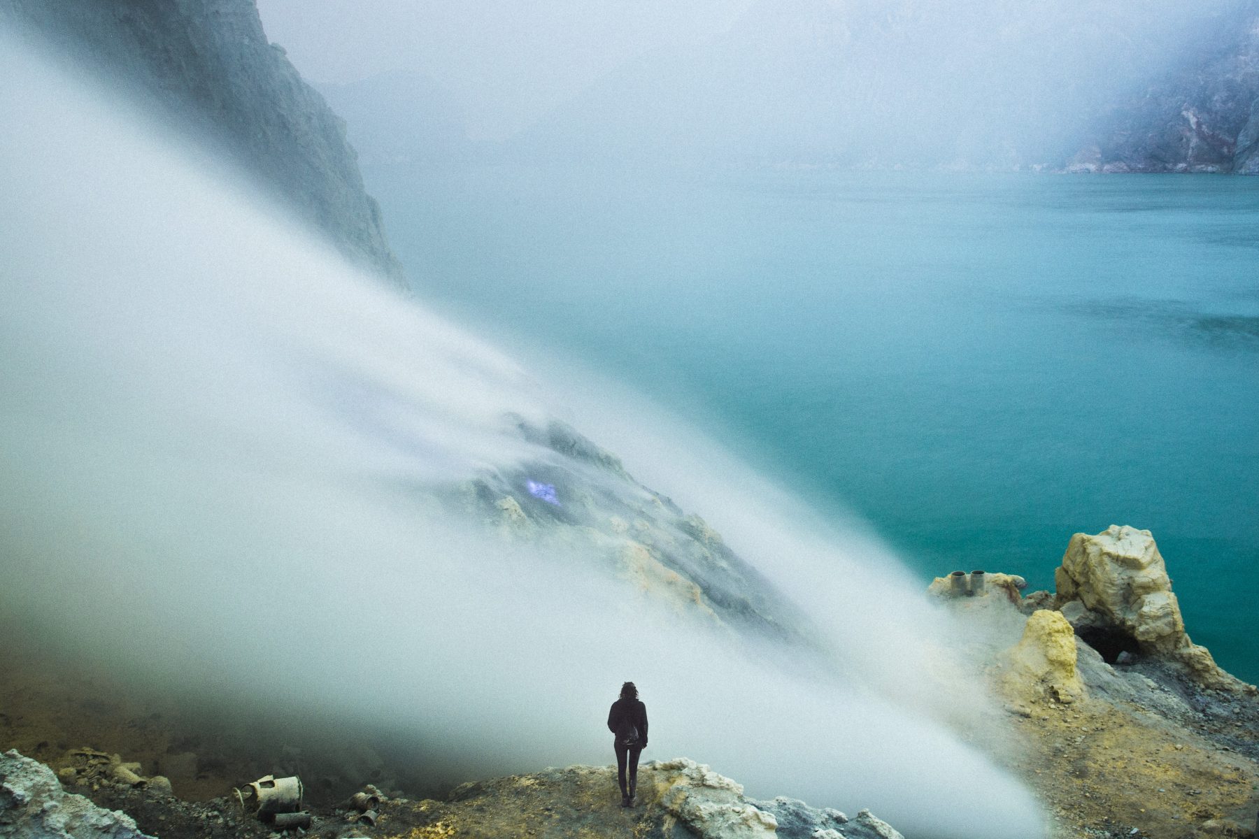 A person stands on a rocky cliff as fog drifts from the water, illuminated by mesmerizing drone light art swirling through the nighttime landscape.