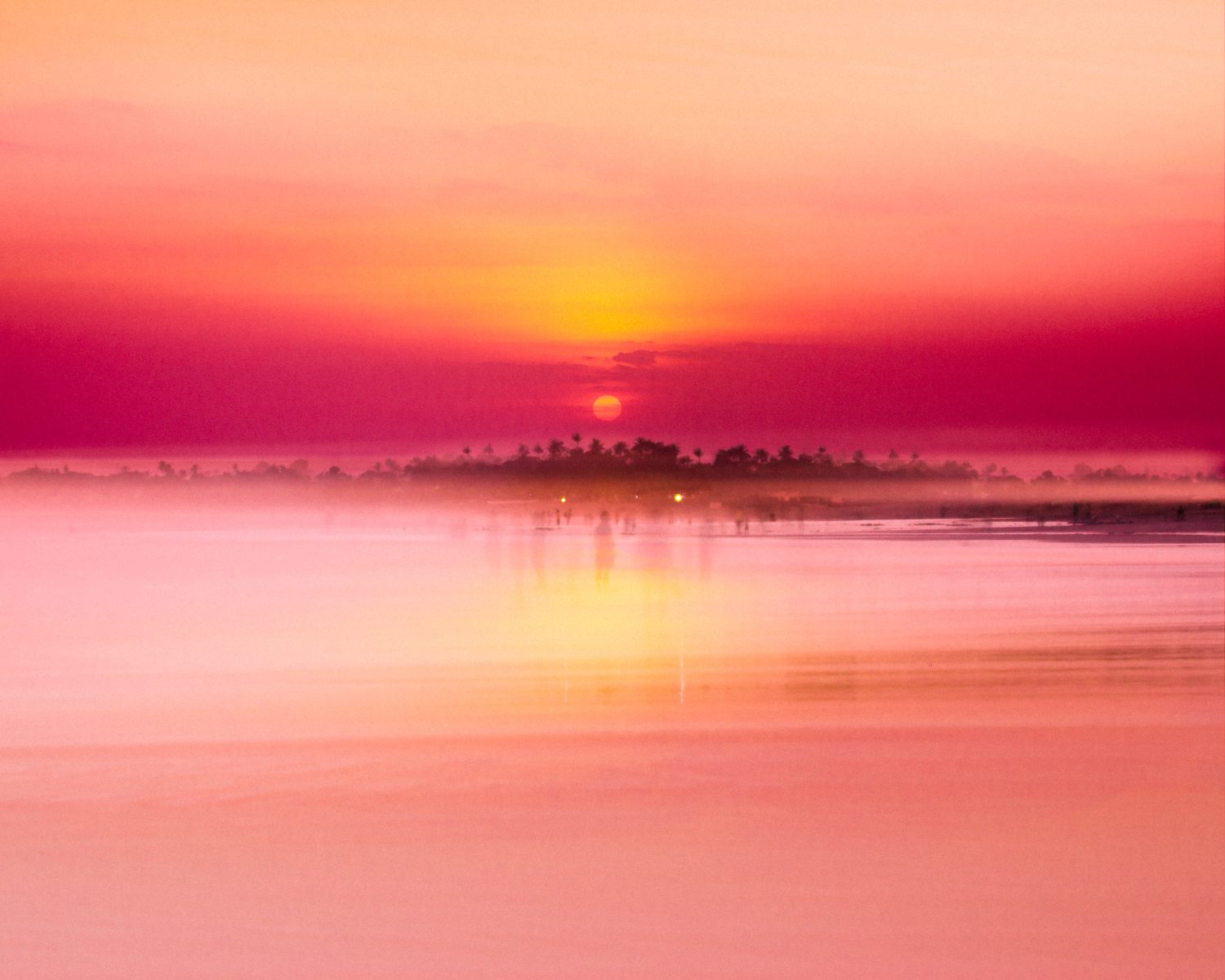 Pink and orange sunset over a calm lake, captured by a photographer, with a distant silhouette of trees and soft mist.