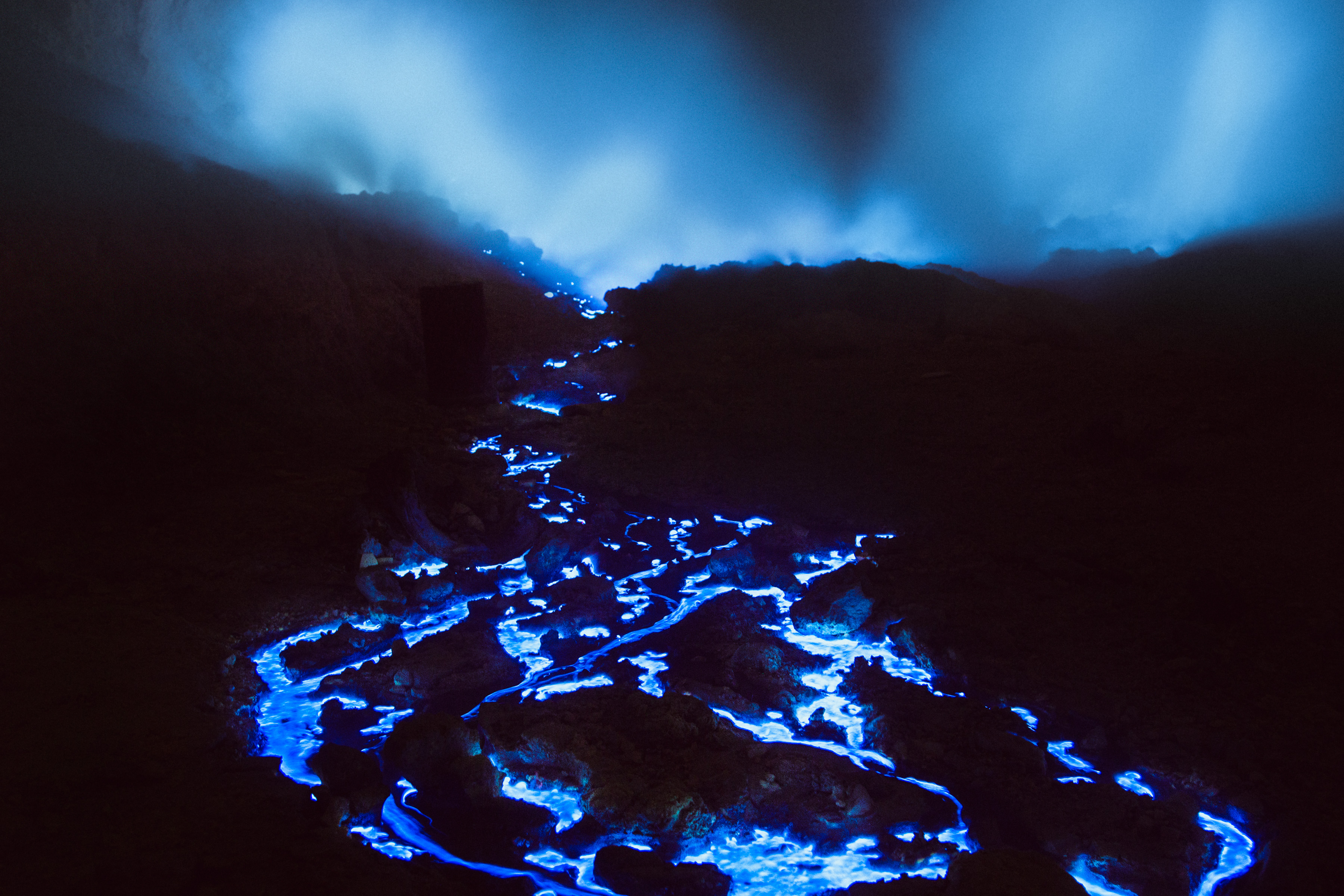 Flowing blue lava glows in the dark, illuminating rocks under a smoky, misty sky—a surreal nighttime landscape photography scene.
