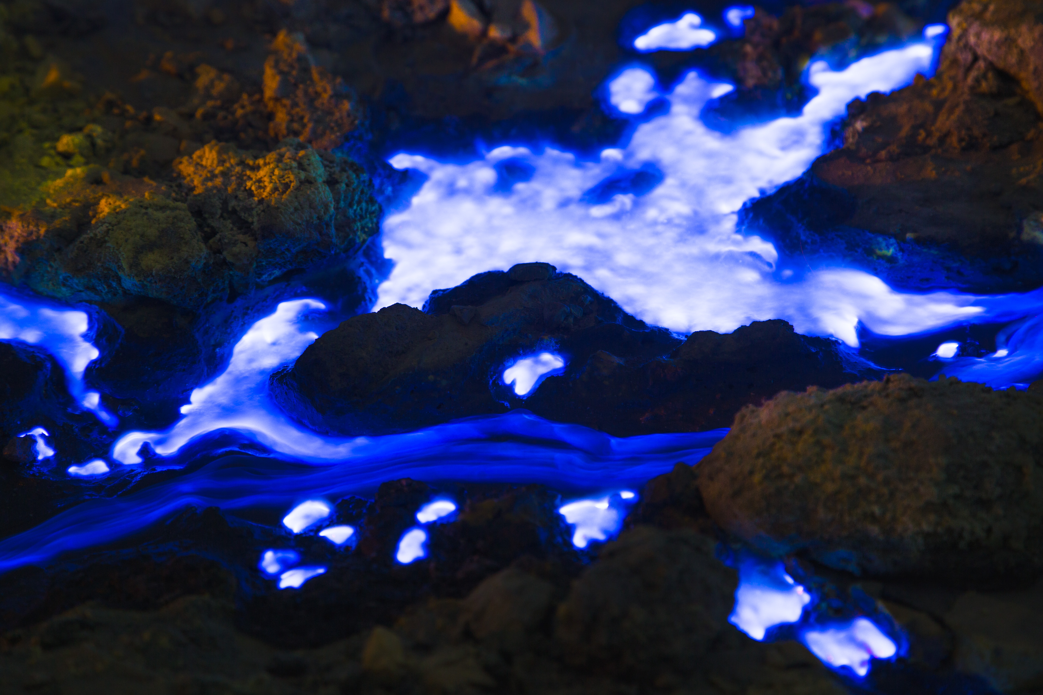 Bright blue glowing liquid flows between dark rocks, creating a striking, futuristic scene reminiscent of nighttime landscape photography.