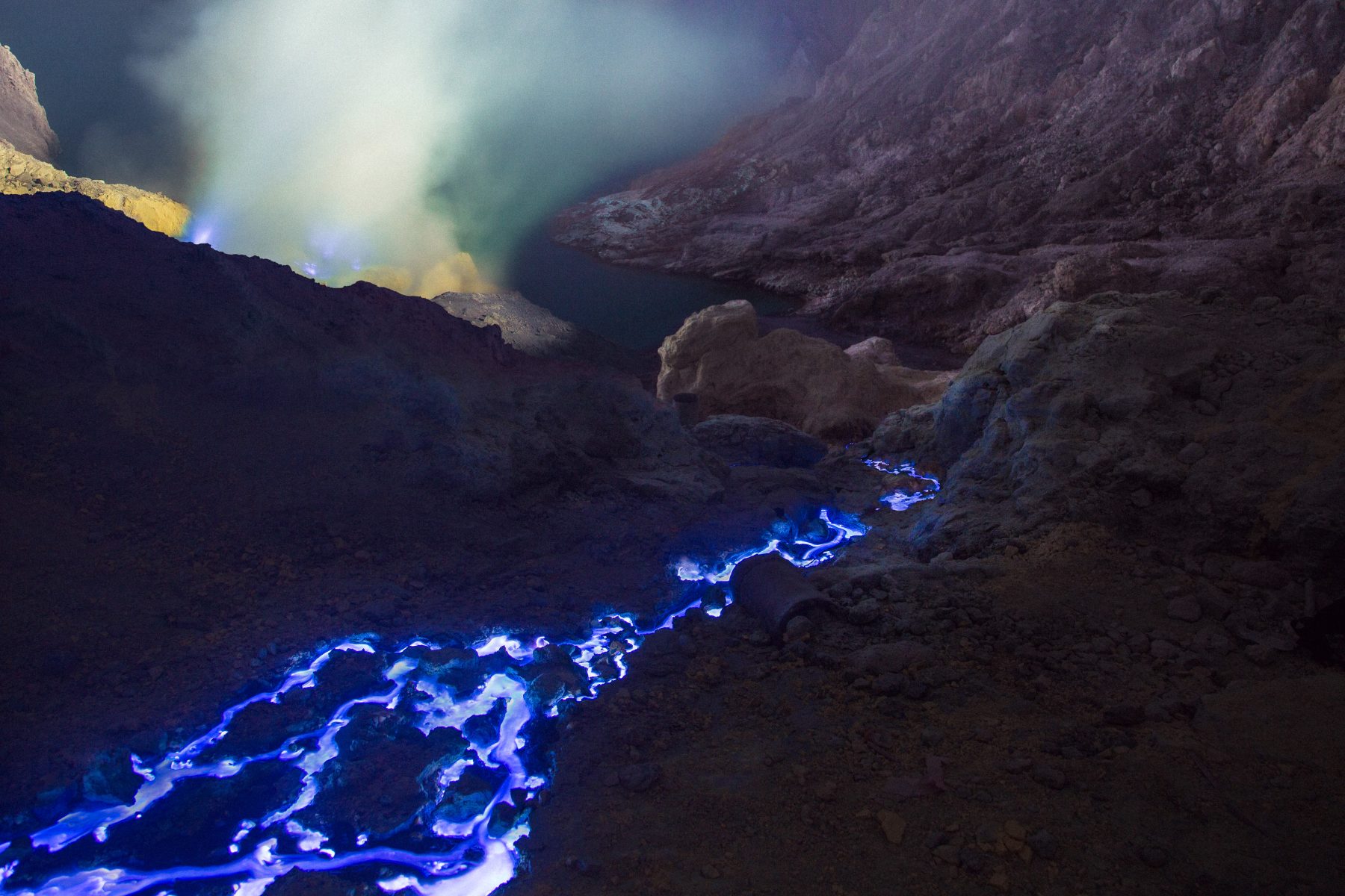 Blue flames and smoke rise from rocky ground at night in a volcanic landscape, captured by a photographer as drone light art dances above the scene.