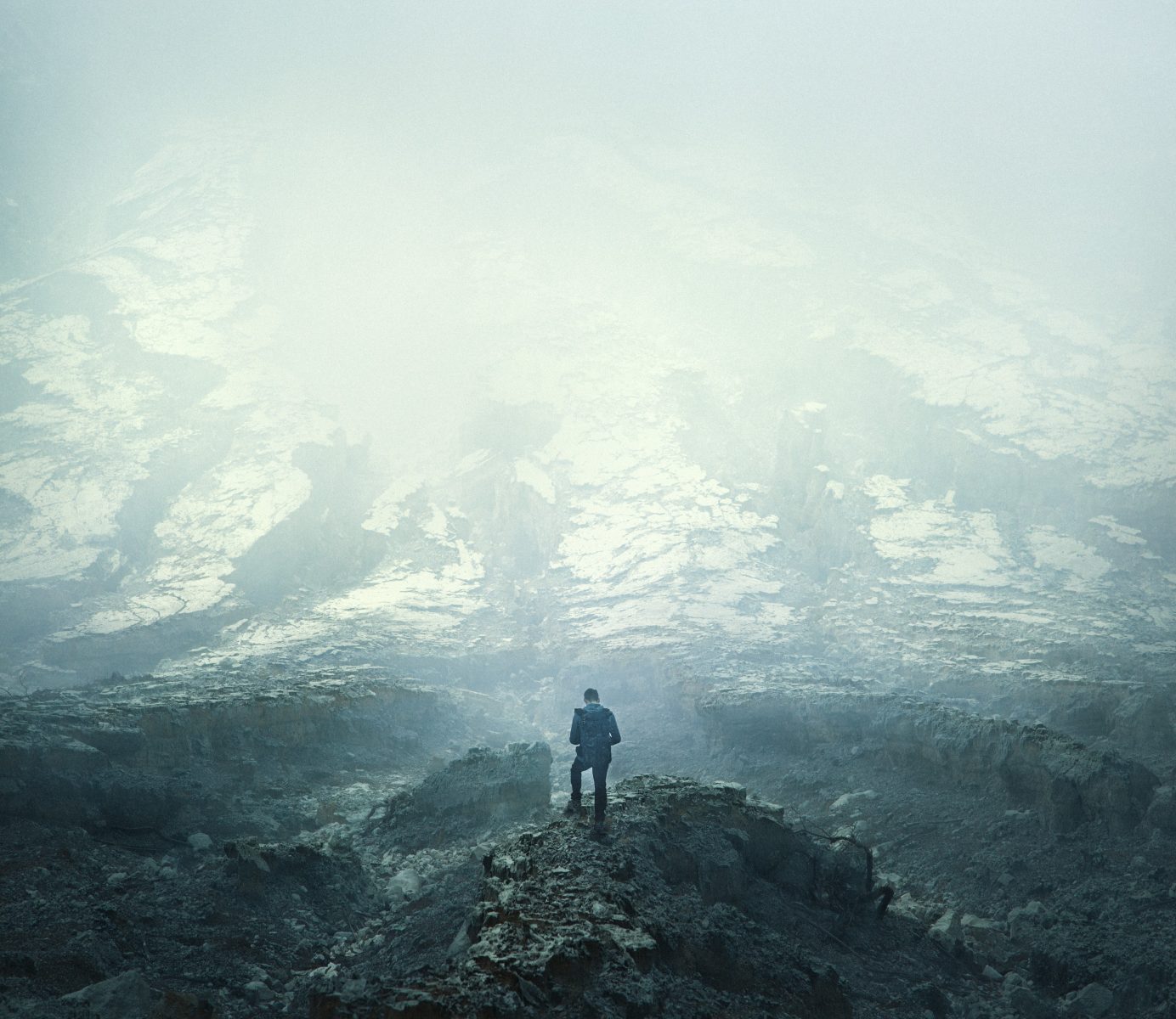 A lone person stands on rocky terrain, surrounded by mist and snow-covered ground under a foggy sky, evoking the mood of fine artist nighttime landscape photography.