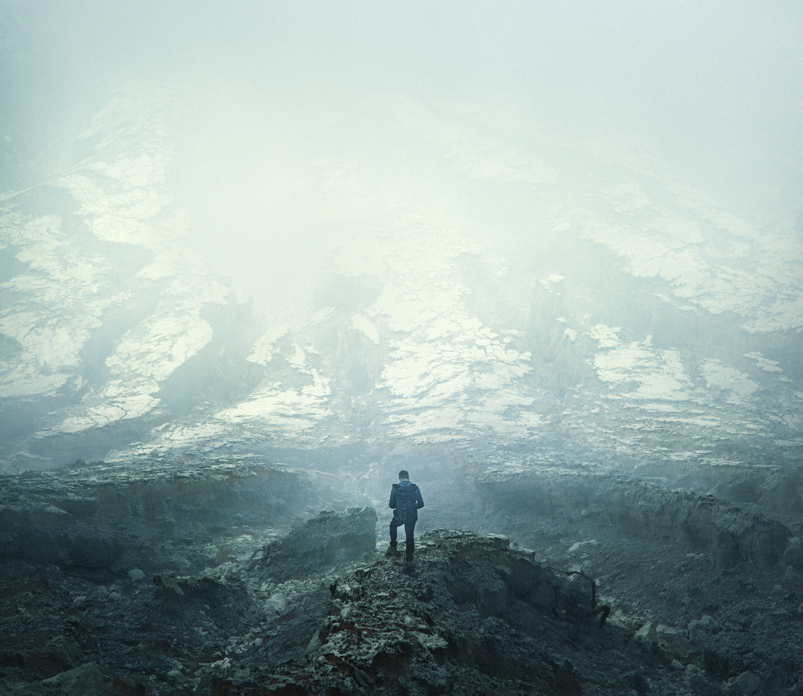 A lone person stands on rocky terrain, surrounded by mist and snow-covered ground under a foggy sky, evoking the mood of fine artist nighttime landscape photography.