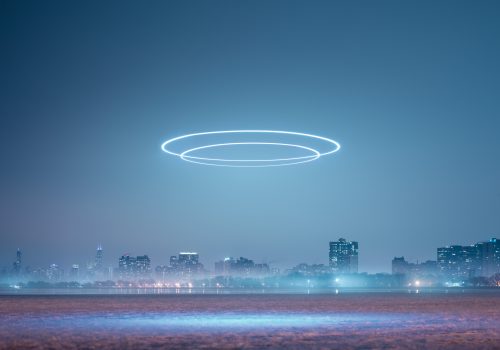 A city skyline at night with glowing drone light art rings illuminating the sky above the buildings.