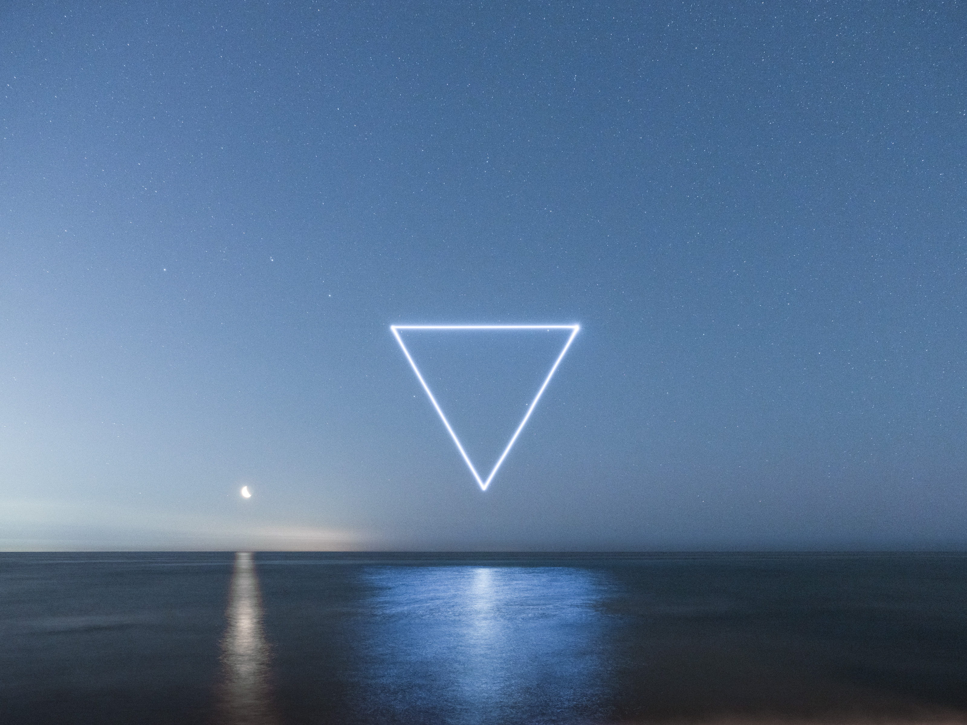 A glowing upside-down triangle, created by drone light art, floats above a calm sea at night under a starry sky and crescent moon.