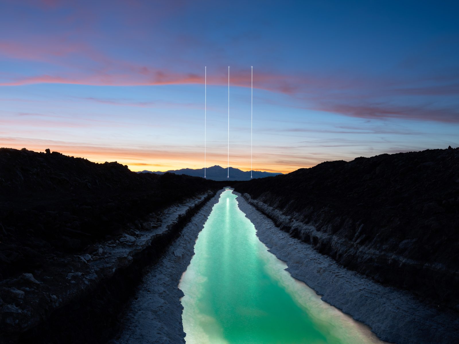 A glowing turquoise canal stretches toward distant mountains under a colorful sunset sky, with white lines above—an ethereal scene captured by a photographer passionate about drone light art.