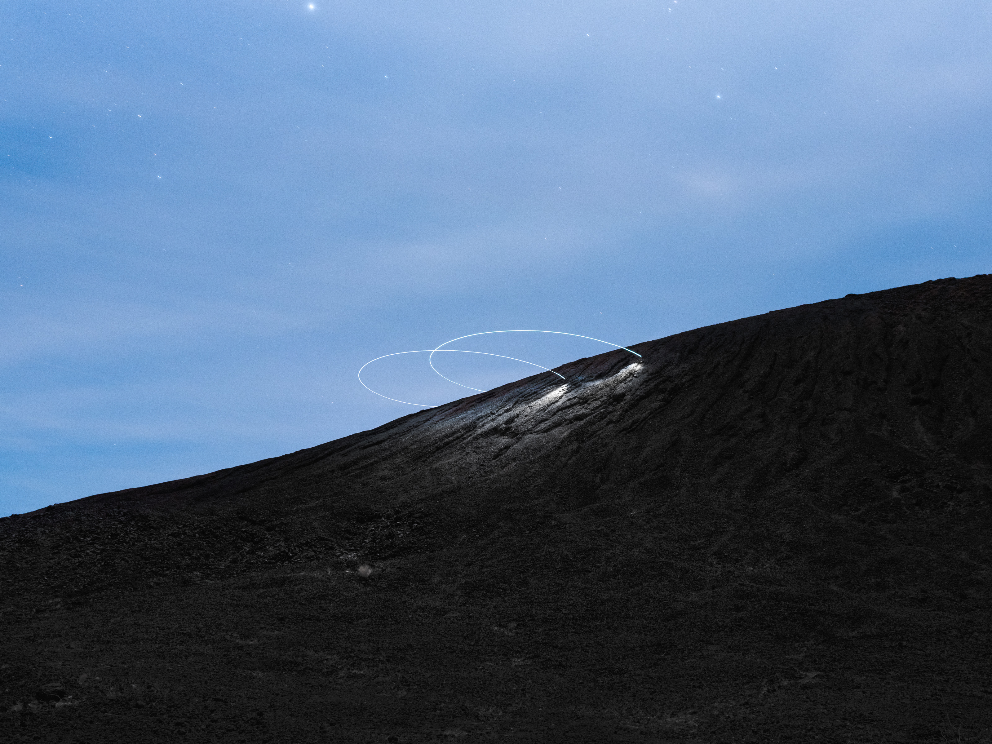 A rocky hillside under a night sky with futuristic drone light art swirling above the ridge.