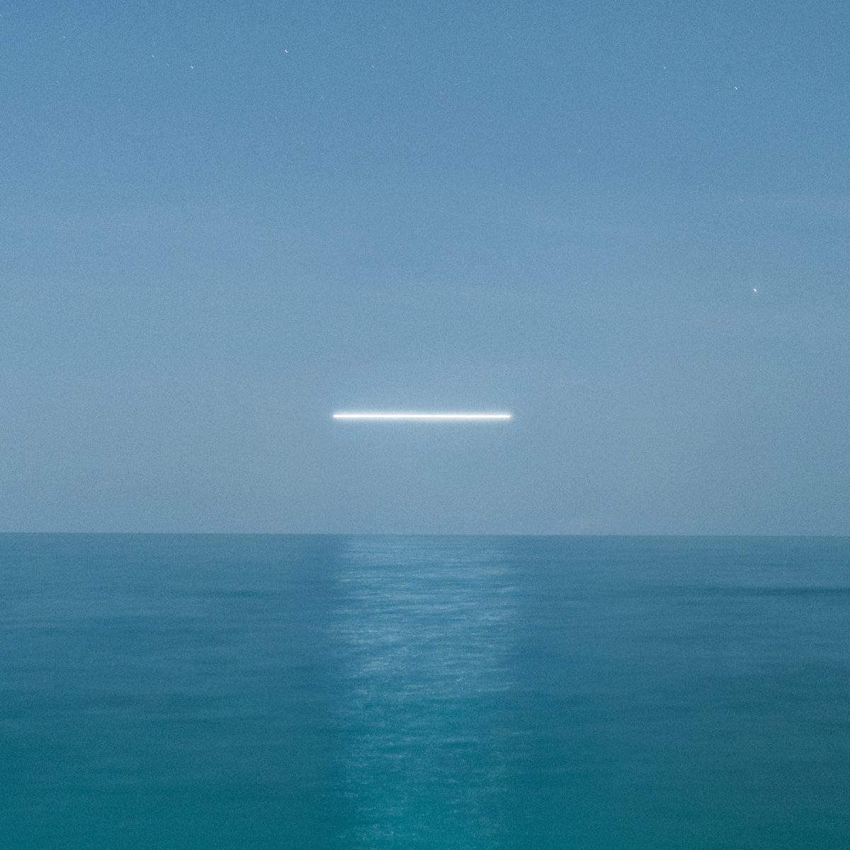 A horizontal white light, created by a drone light art photographer, hovers above calm blue ocean water under a clear sky at night.