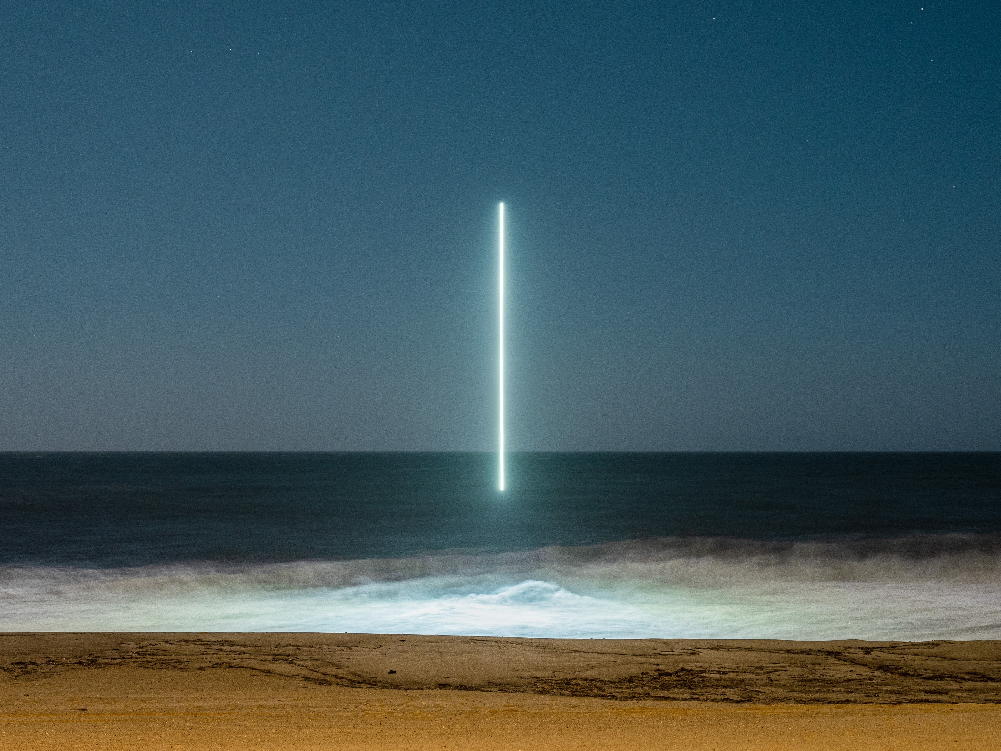 A bright vertical line of light hovers over the ocean at night, seen from a sandy beach—a mesmerizing scene for any photographer passionate about nighttime landscape photography.