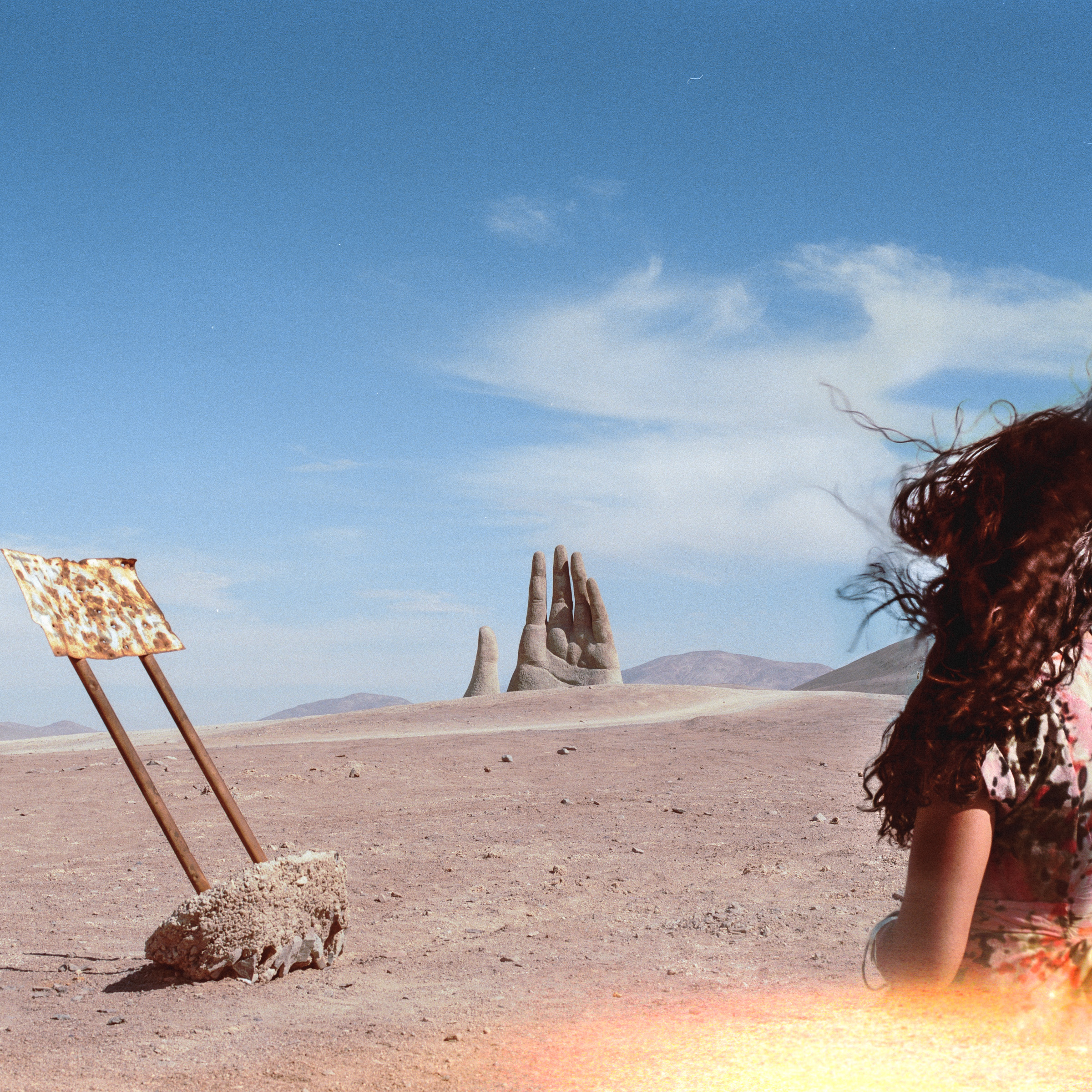 A photographer faces a giant hand sculpture rising from a desert, with a rusty sign on the left.