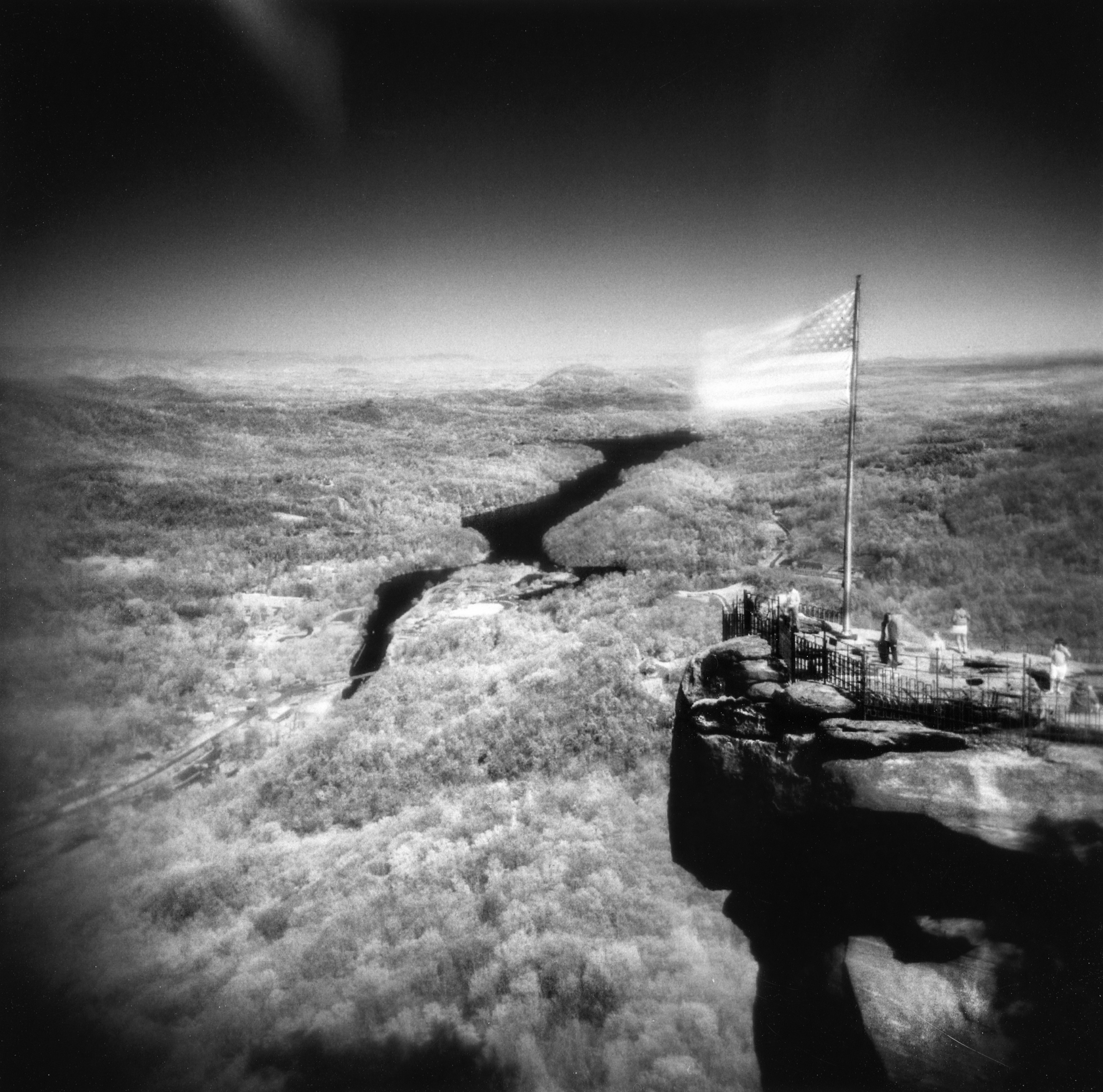 Black and white photo of a scenic overlook with an American flag, river, and distant hills, captured by a fine artist with a photographer’s eye for dramatic compositions.