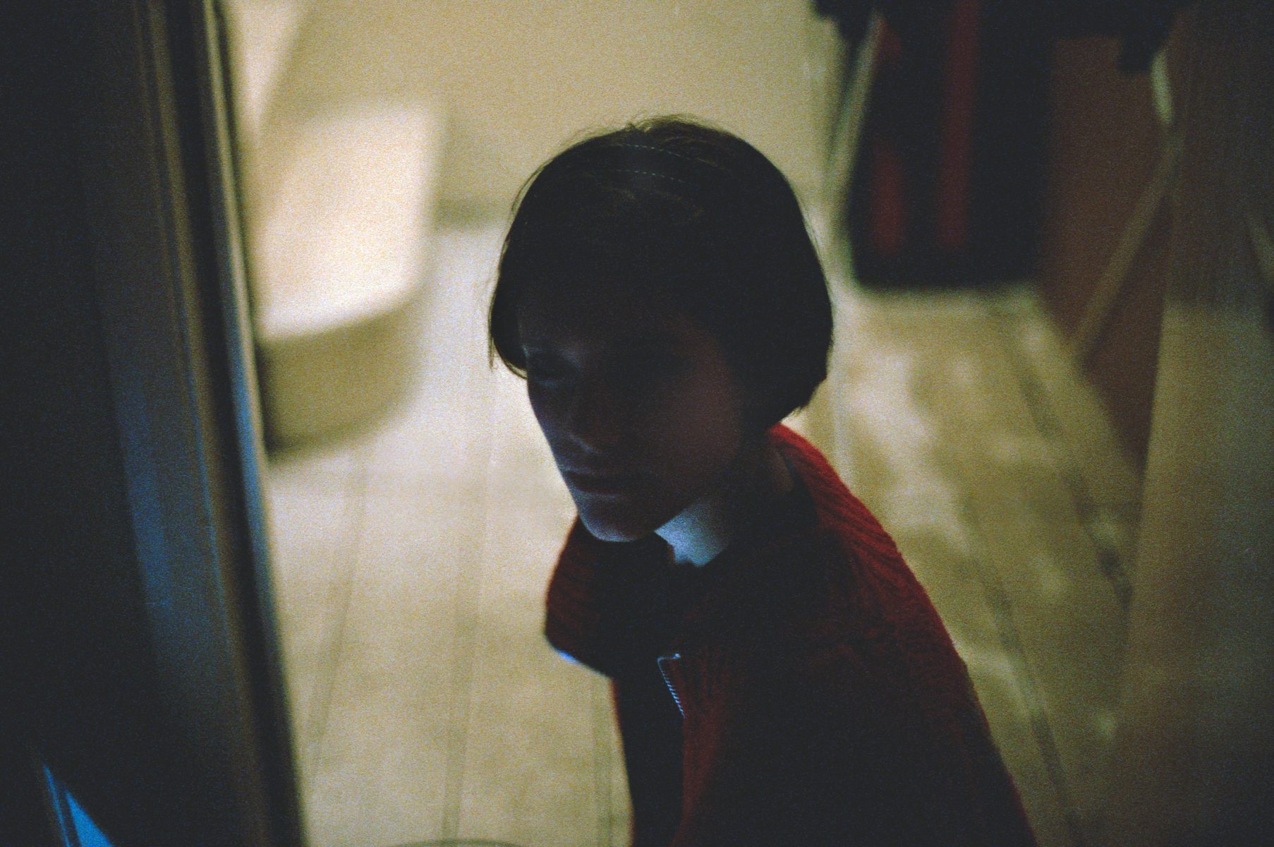 A person with short dark hair in a red sweater sits in a dimly lit room, looking toward the camera, as if posing for a futuristic nighttime landscape photography session.
