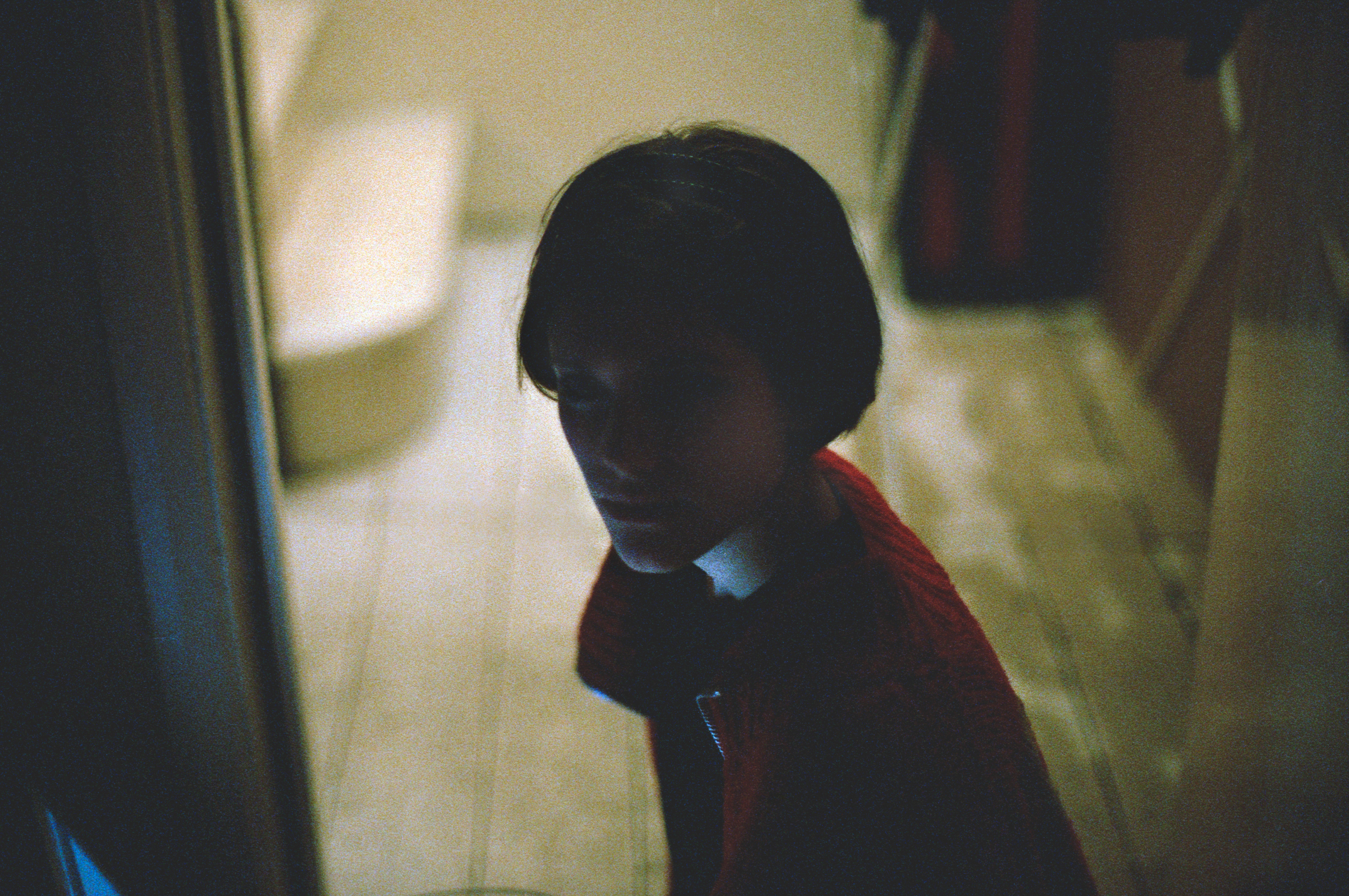 A person with short dark hair in a red sweater sits in a dimly lit room, looking toward the camera, as if posing for a futuristic nighttime landscape photography session.
