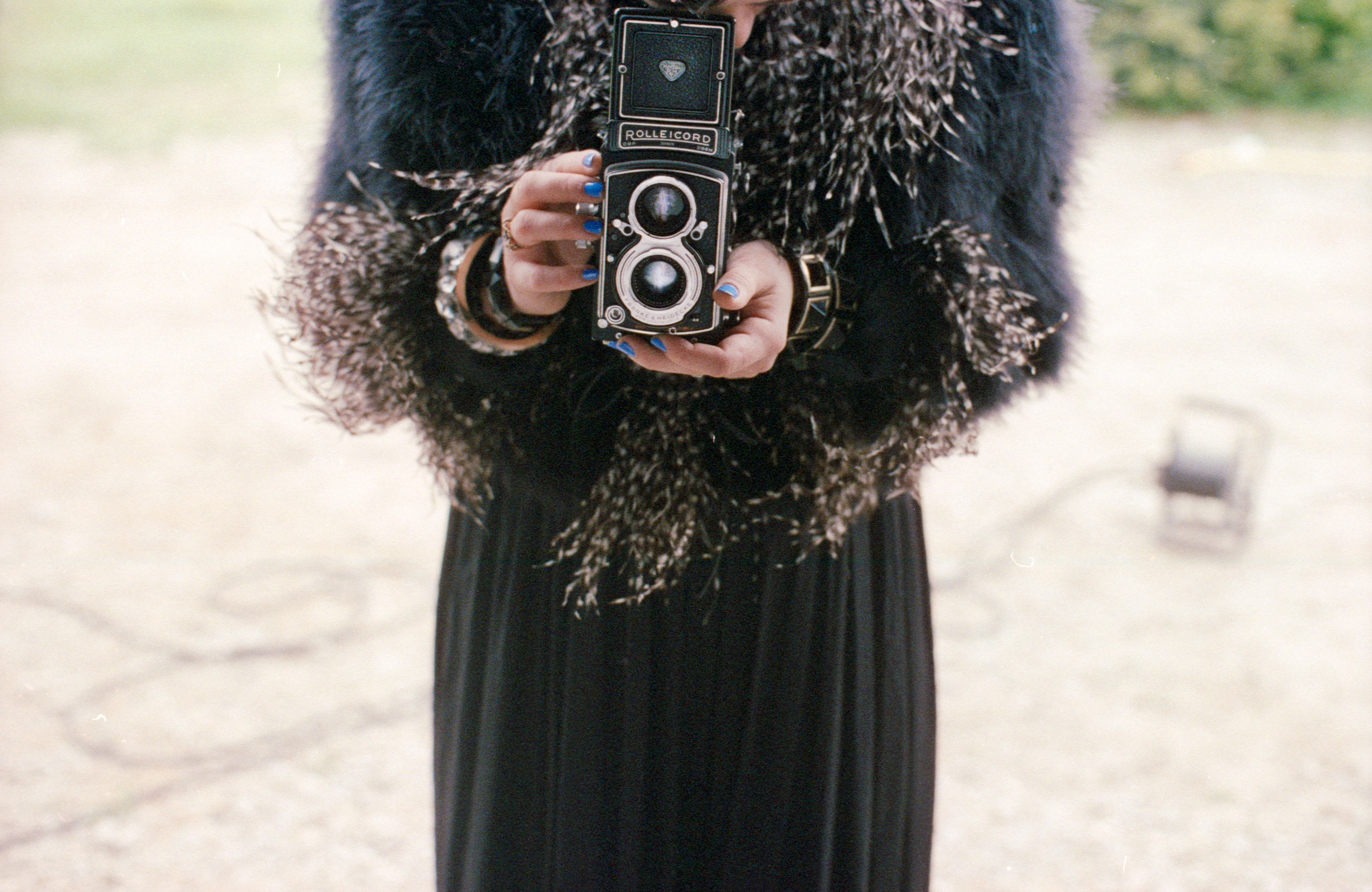 A person in a black outfit holding a vintage twin-lens camera, with blue nail polish visible, stands ready to capture futuristic nighttime landscape photography.