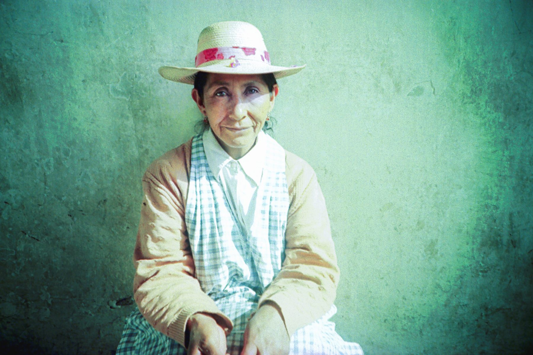 Woman in a straw hat and checked apron sits against a textured, pale green wall, smiling gently—like a photographer pausing to reflect before capturing drone light art or nighttime landscape photography.