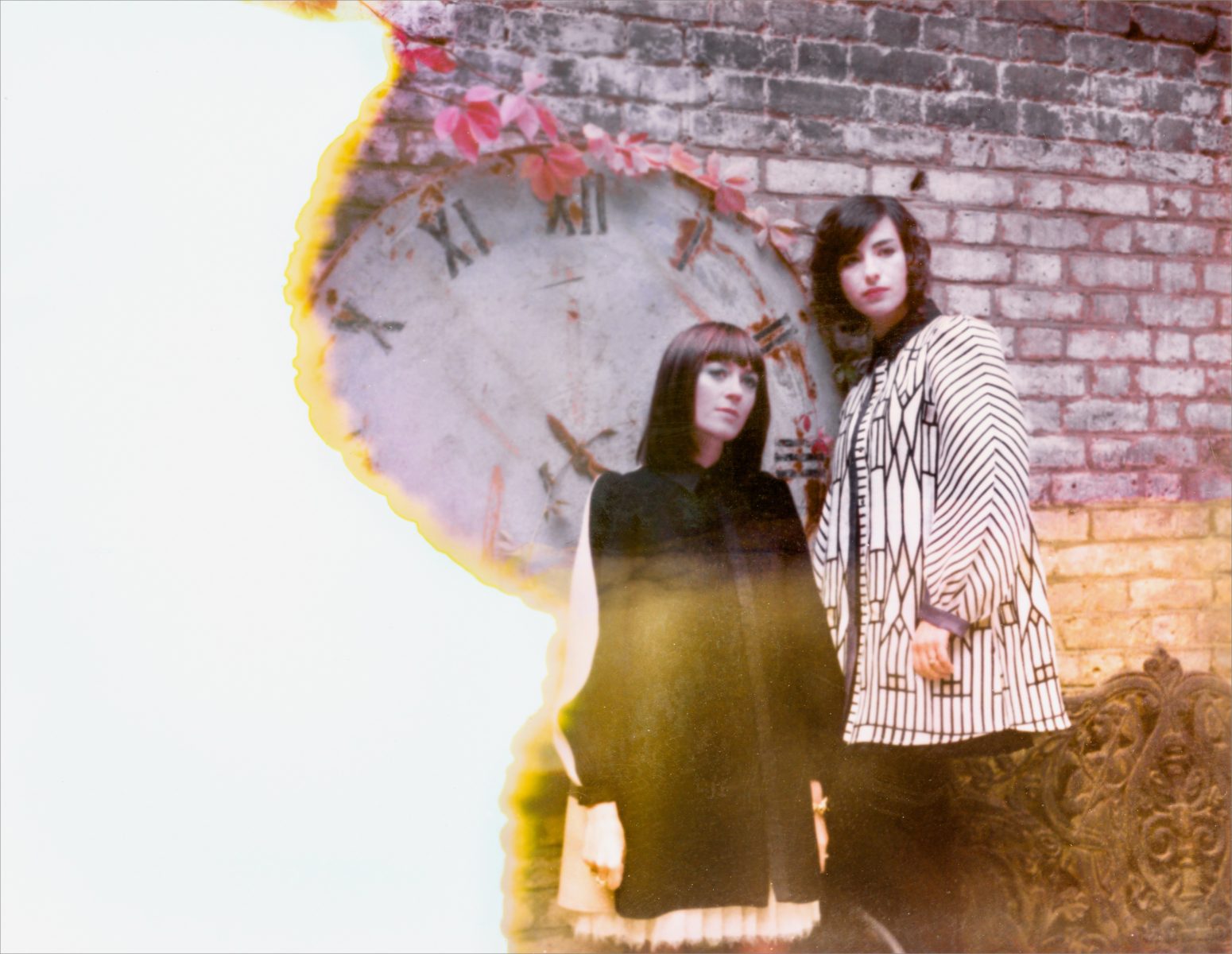 Two women stand by a brick wall and faded clock, with the photo partially burned on the left side—a striking composition by a skilled photographer.
