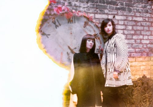 Two women stand by a brick wall and large clock; the photo, partly damaged with a burnt effect on the left side, recalls the creative vision of a fine artist experimenting with texture and time.